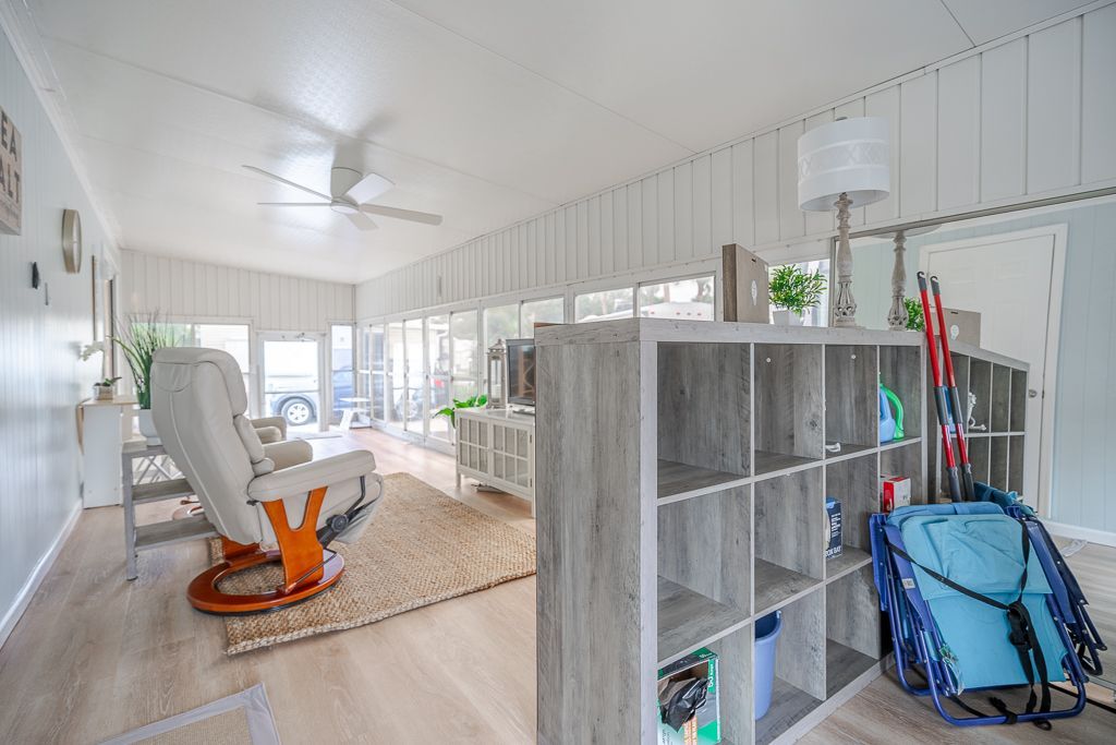 A bright sunroom with light wood flooring, a white recliner on a textured rug, and a gray open shelving unit.