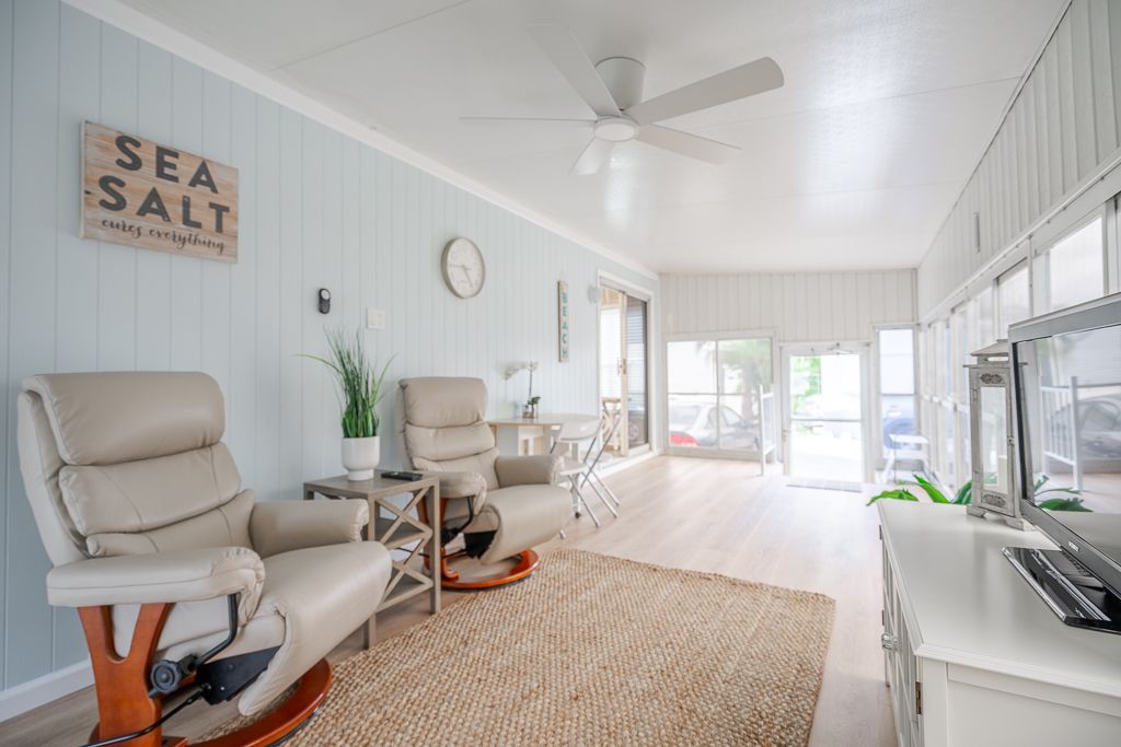 A light-filled sunroom features two tan recliner chairs, a woven area rug, and soft blue vertical paneled walls.