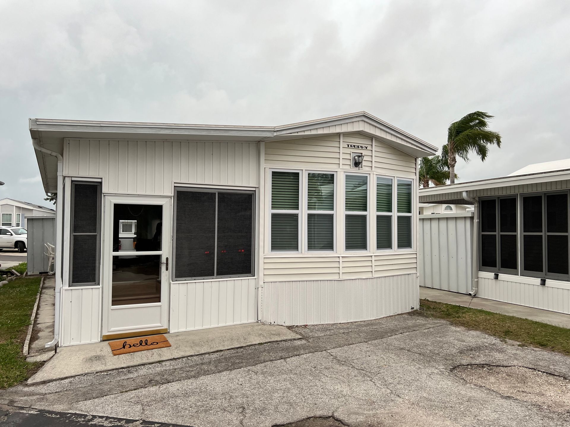 A white manufactured home with a bay window, a glass door, and a gravel driveway under an overcast sky.