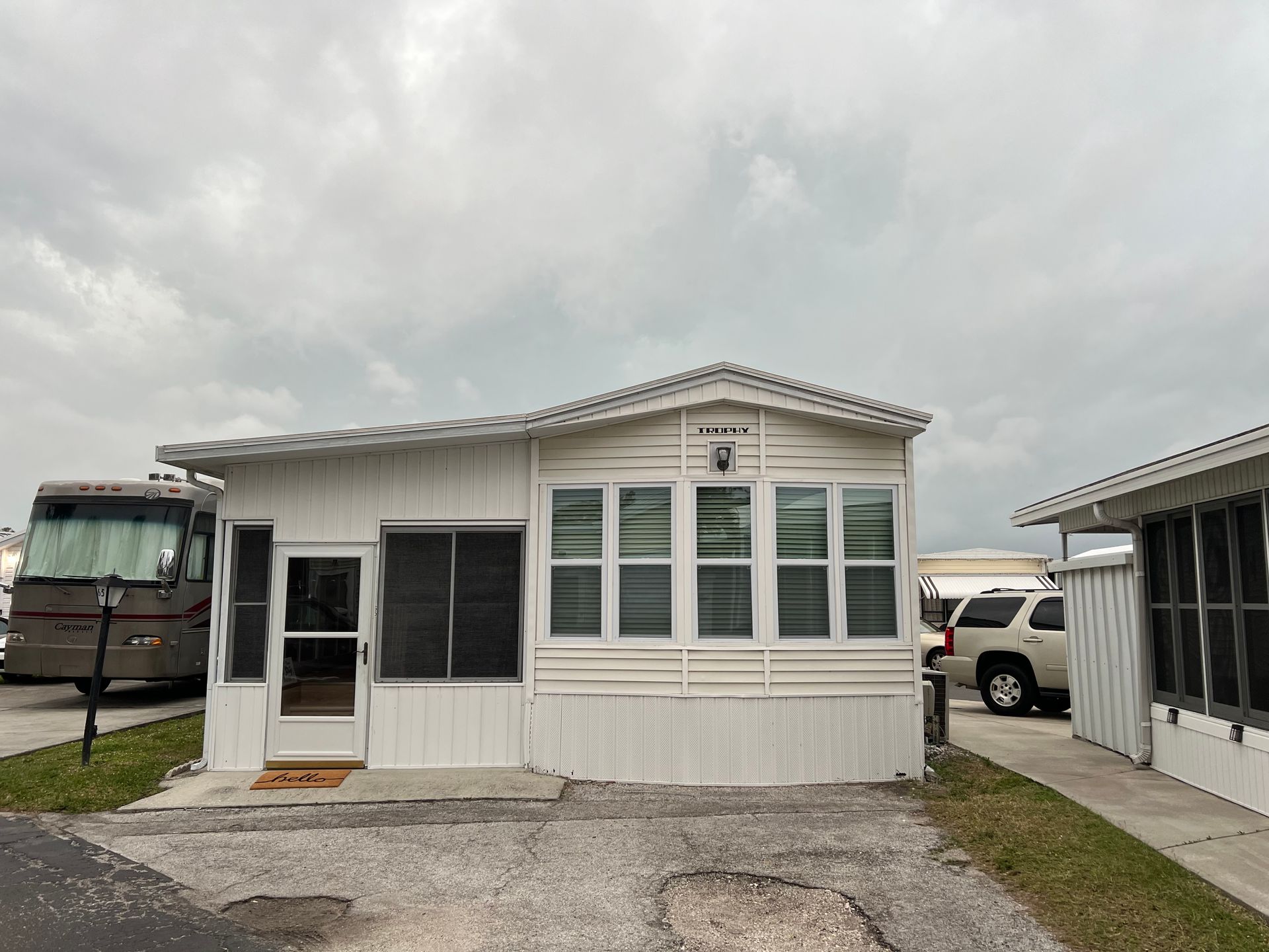 A single-story, white manufactured home with a gravel driveway, parked RV to the left, and cloudy sky overhead.
