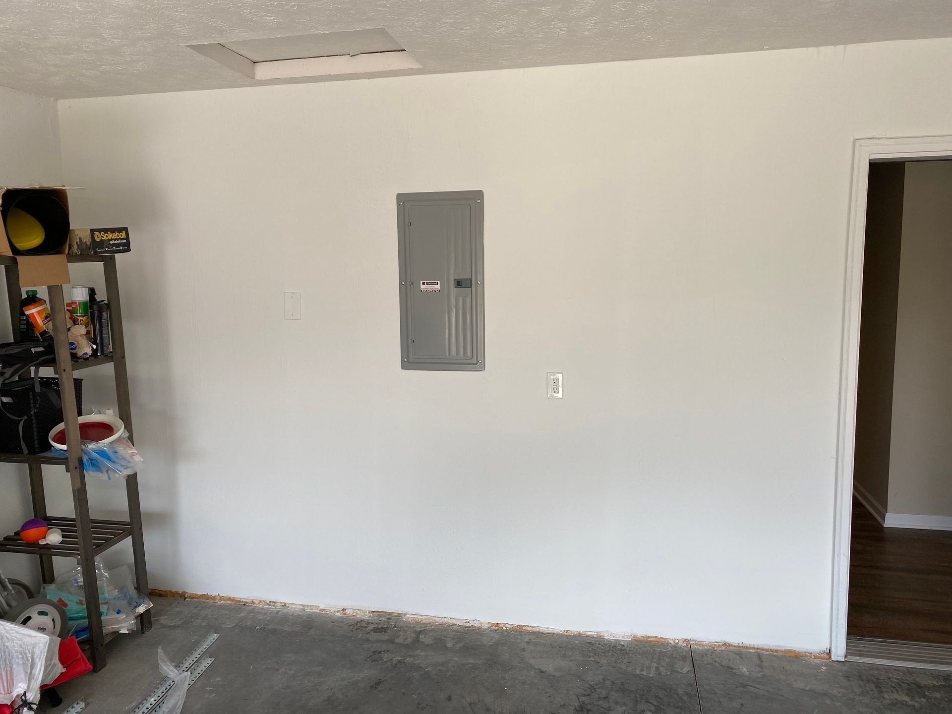 A gray electrical panel mounted on a white wall next to an open doorway, with a metal shelving unit on the left.
