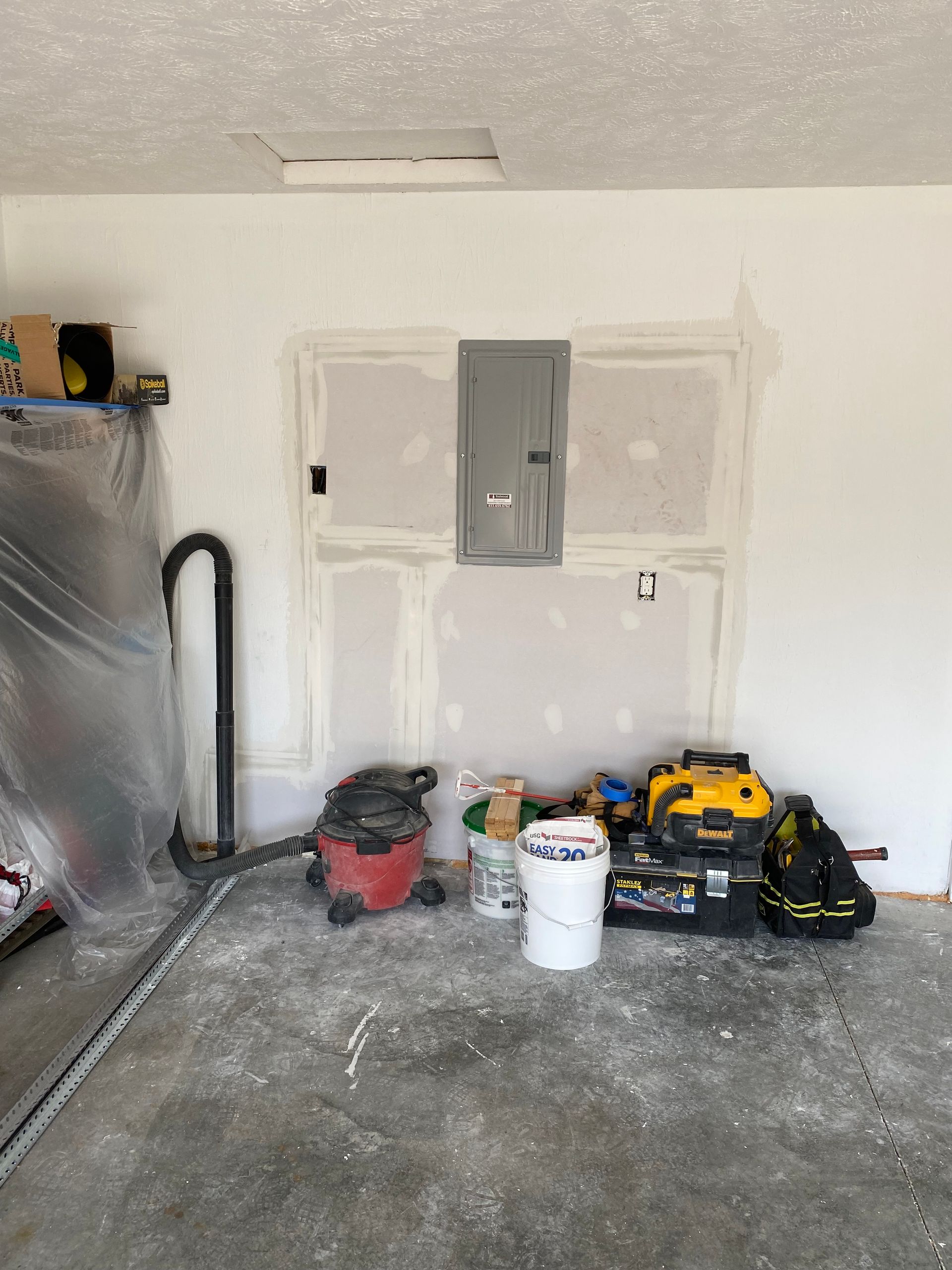 A gray electrical panel installed on a freshly patched drywall wall in a garage, with tools and buckets on the floor.