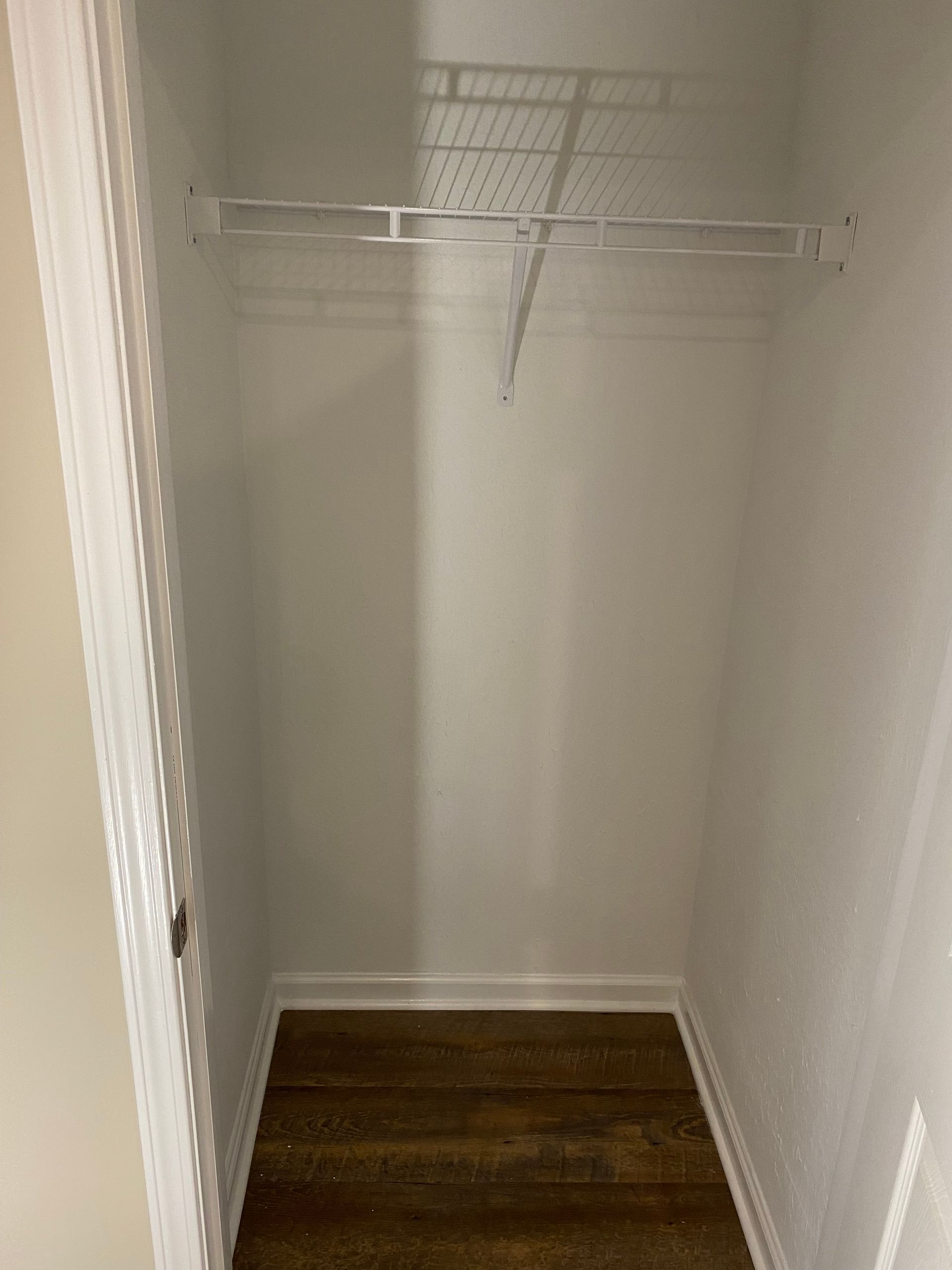 An empty walk-in closet with white walls, white wire shelving, and dark wood-look flooring.