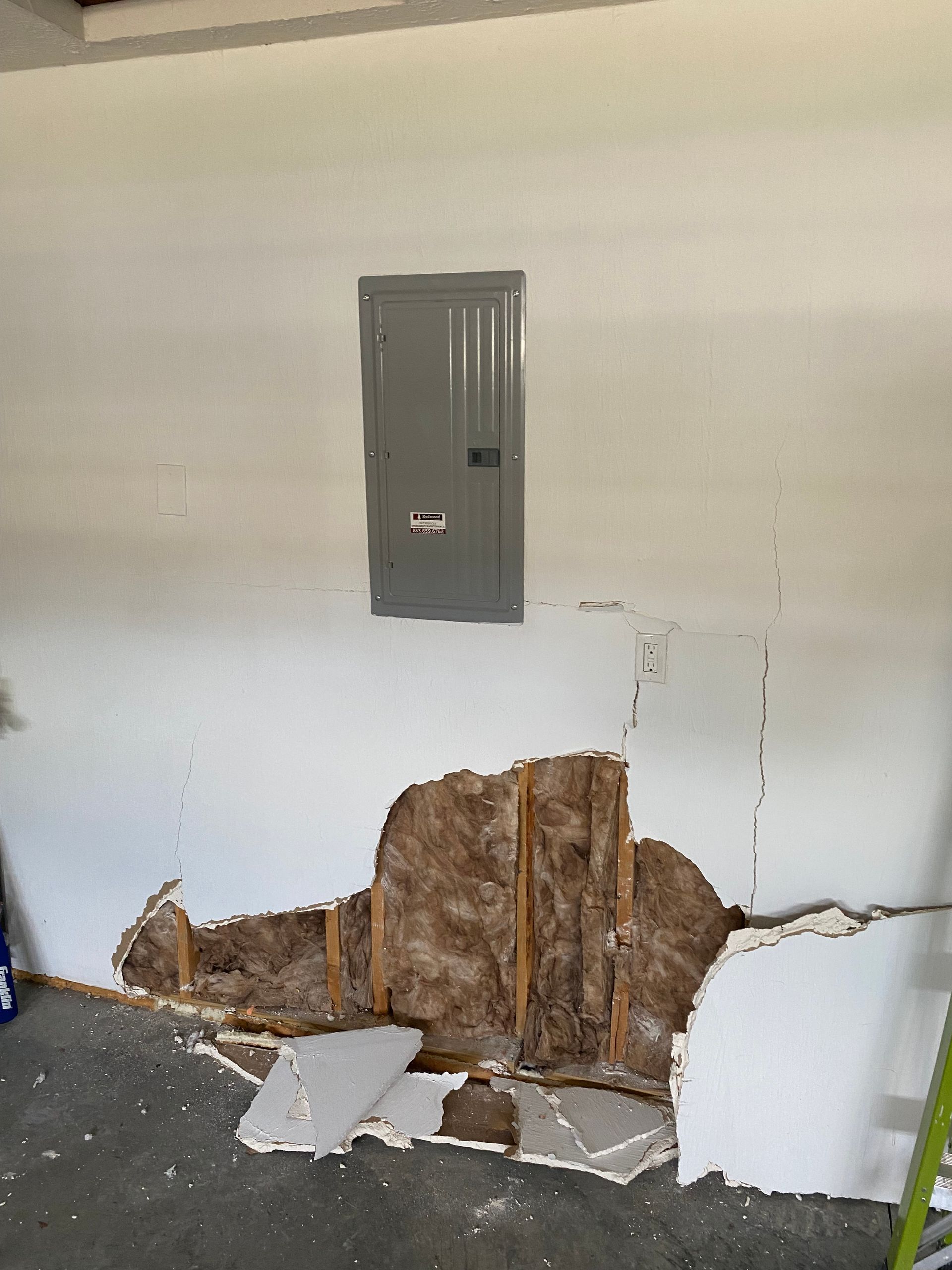 A wall with a gray electrical panel and a large section of damaged drywall exposing wood studs and insulation below.