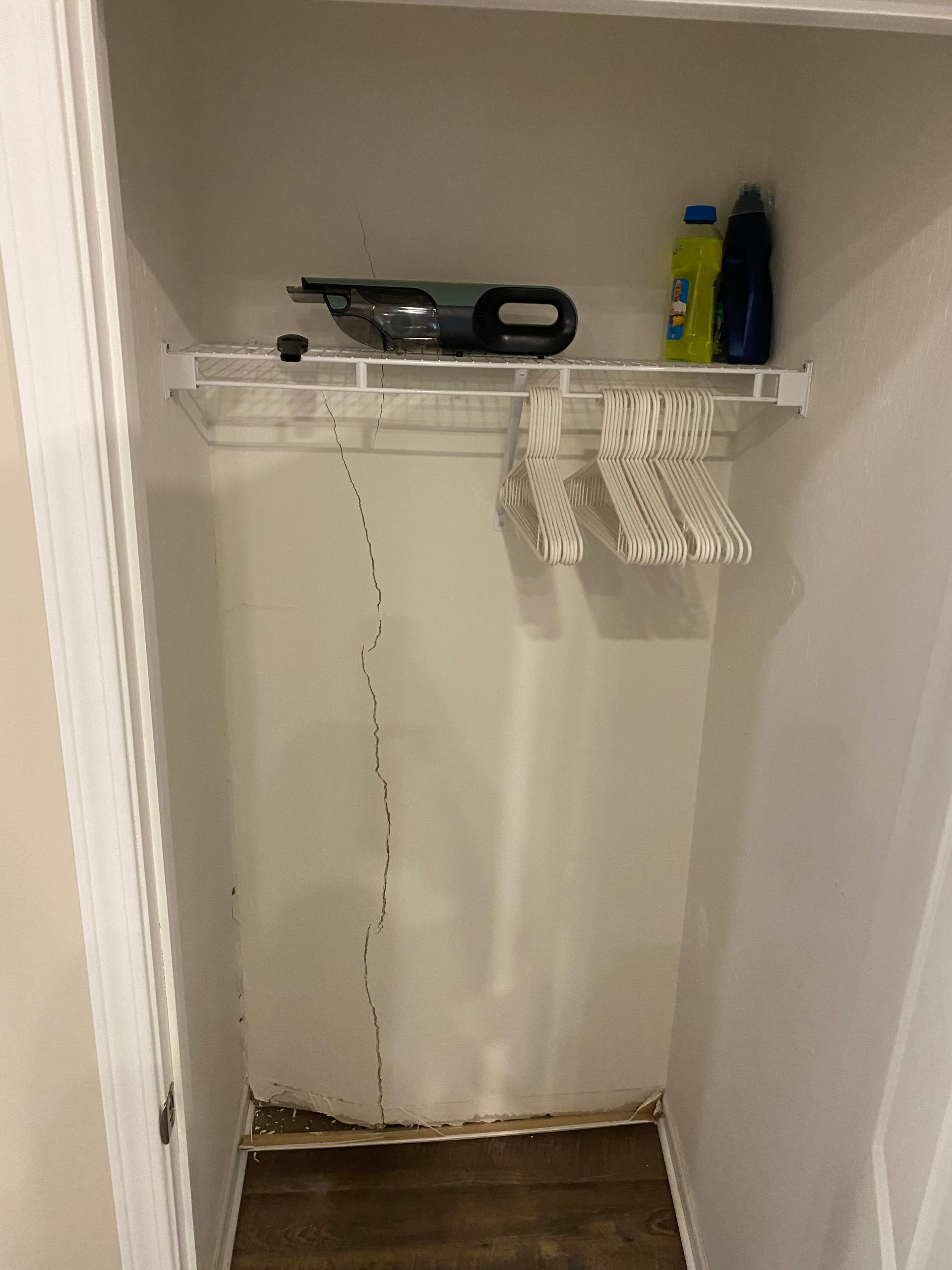 A closet with a long vertical crack in the wall, a wire shelf holding a vacuum and cleaners, and empty clothes hangers.