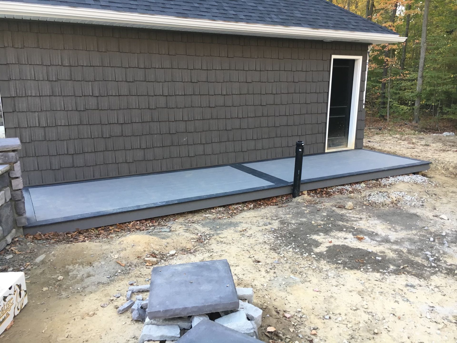 A gray building with dark shingles and a newly installed gray porch deck sits on a dirt lot with stacked stone tiles nearby.
