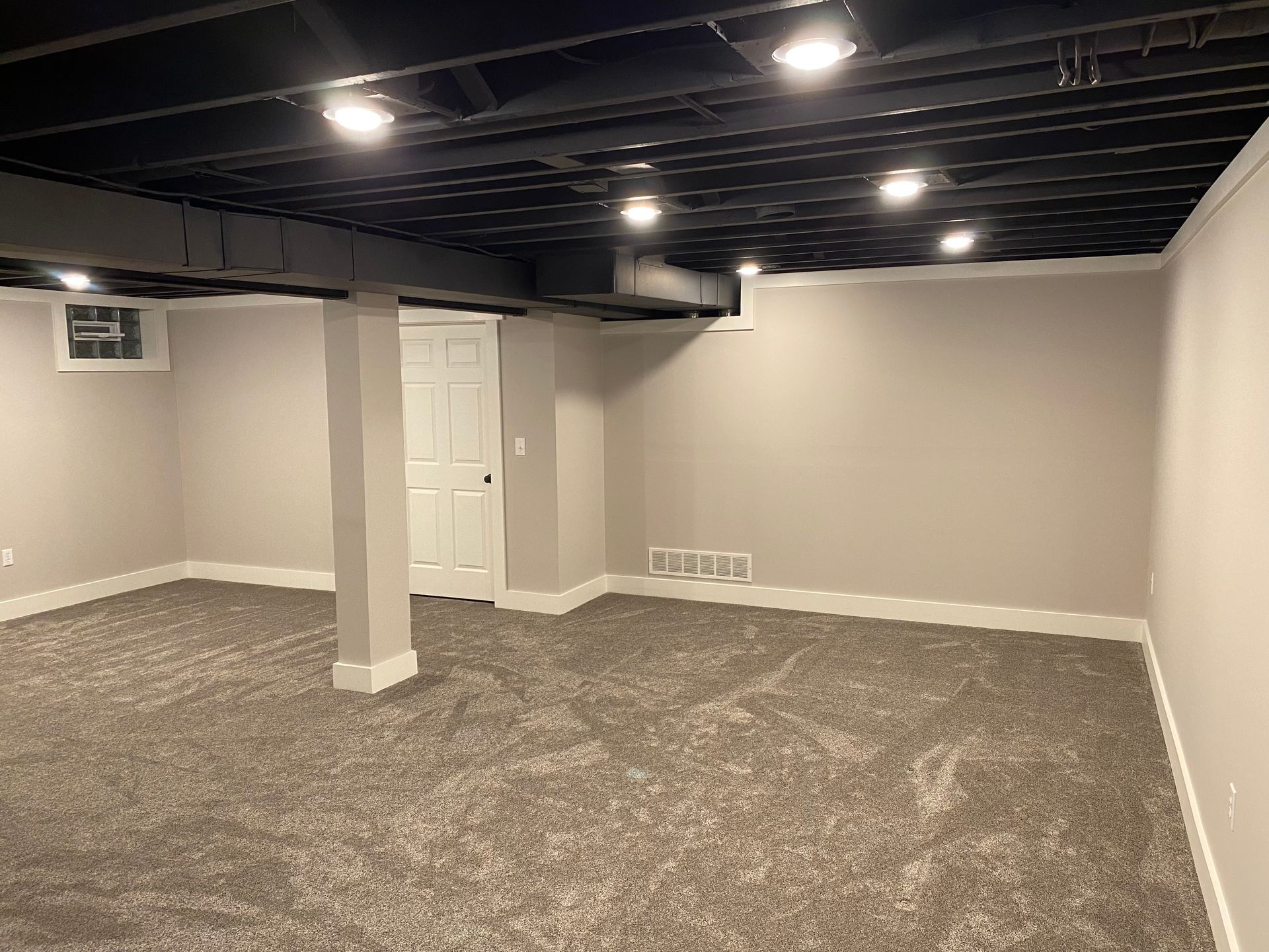 A finished basement with neutral gray carpet, light gray walls, a white door, a support pillar, and a black exposed ceiling.
