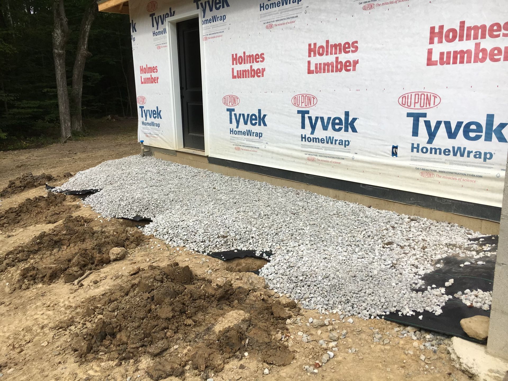 Gravel laid over landscape fabric against the Tyvek-wrapped wall of a house under construction.