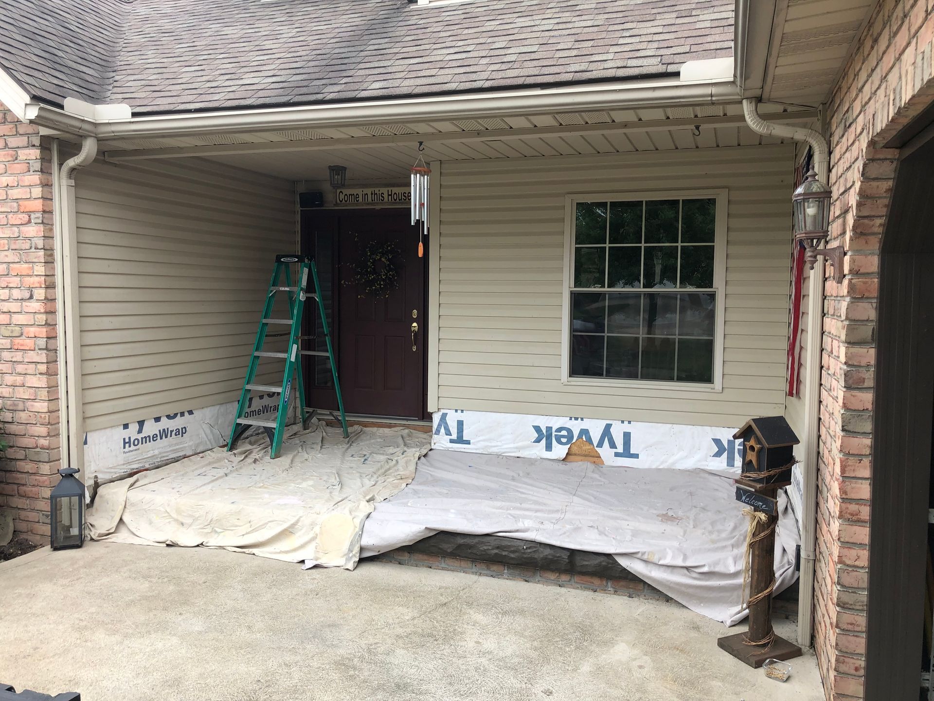 A residential porch under renovation with Tyvek wrap on the walls, a drop cloth on the floor, and a ladder by the front door.