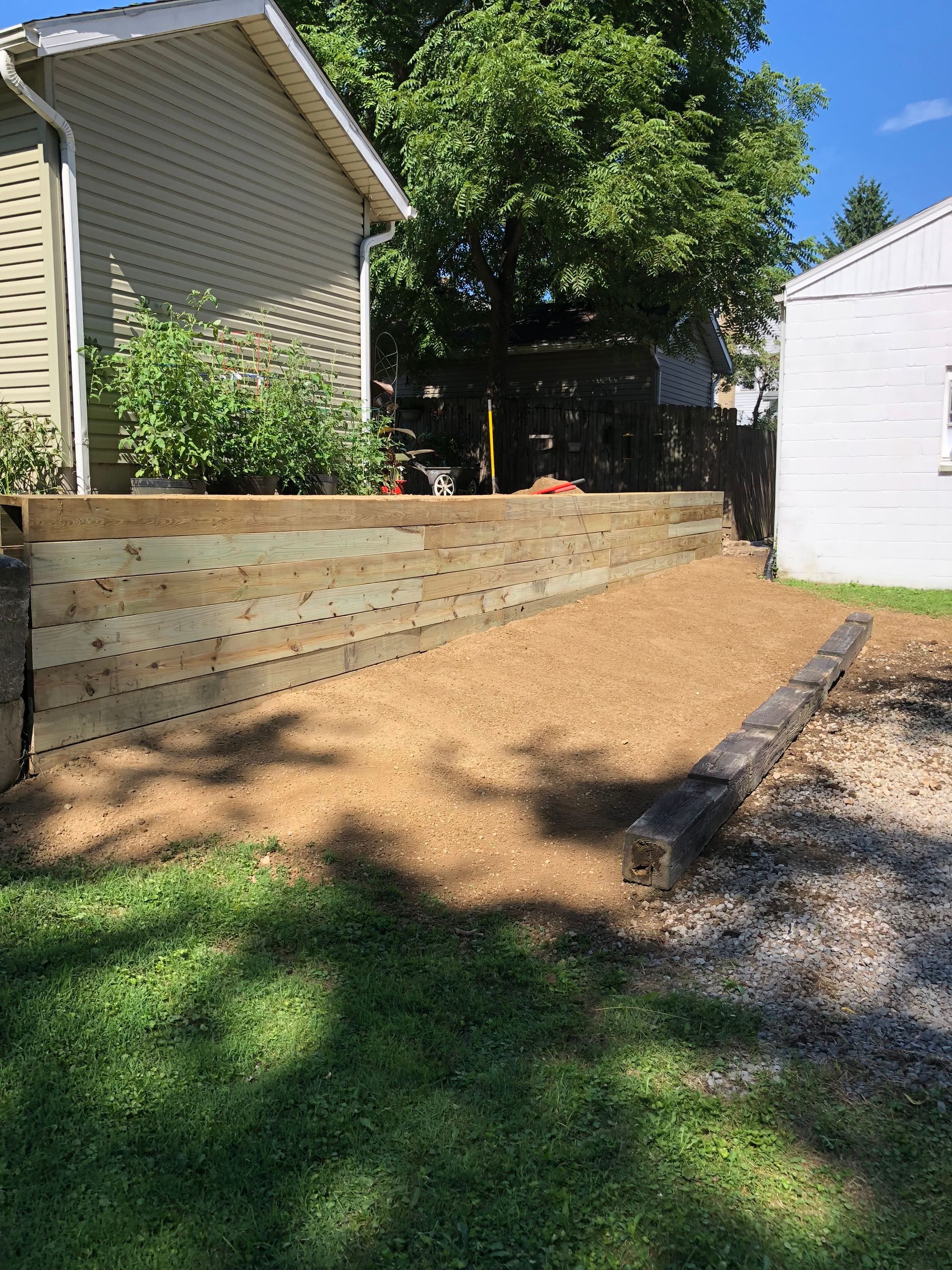 A new light-colored wooden retaining wall sits in a yard with a wood-chipped area and a single old landscape timber.