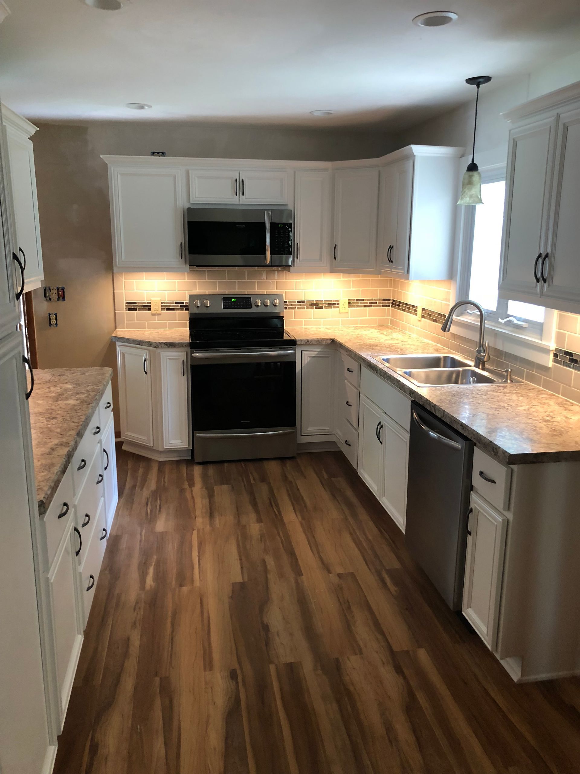 A modern kitchen with white cabinets, granite countertops, stainless steel appliances, and wood-patterned flooring.
