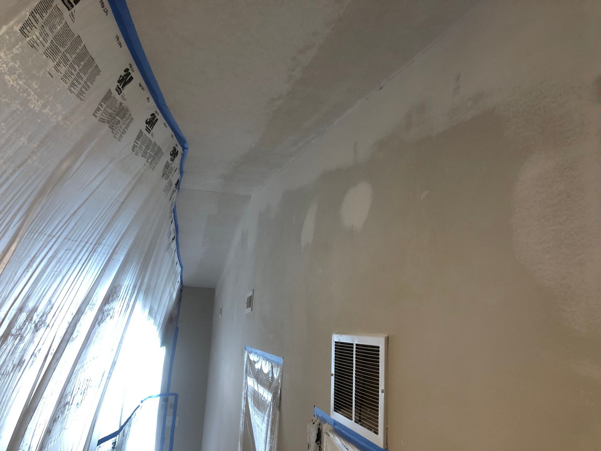 An indoor wall under renovation with patched drywall, blue painter's tape, and a plastic-covered window frame.