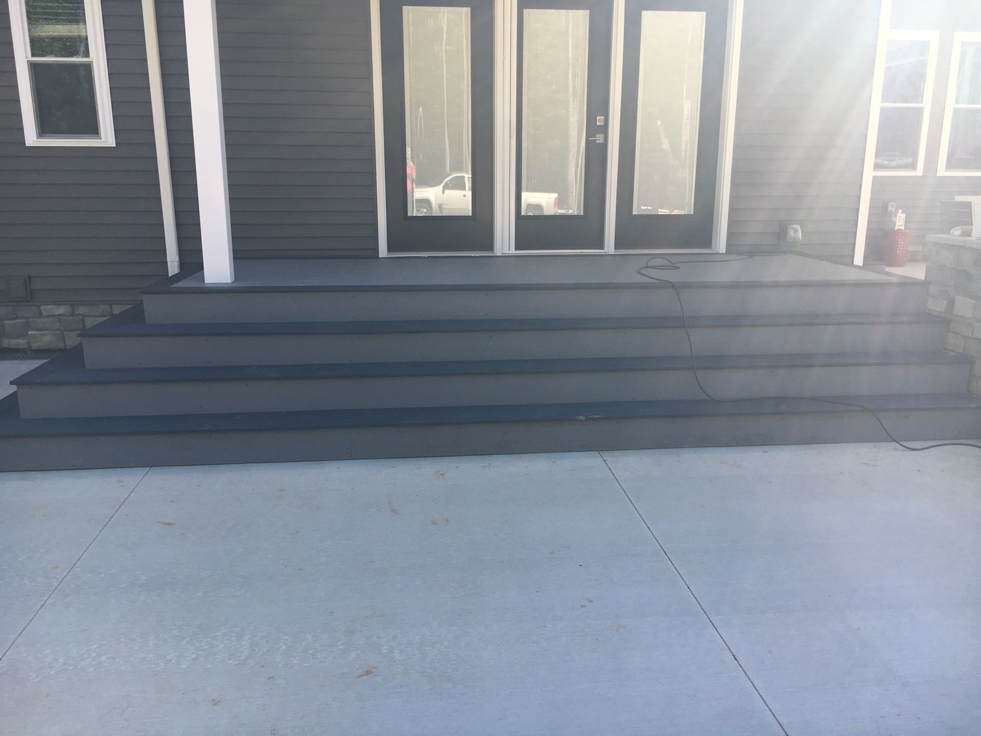 A set of four dark gray steps leading up to double glass doors on a house exterior with light gray concrete in front.