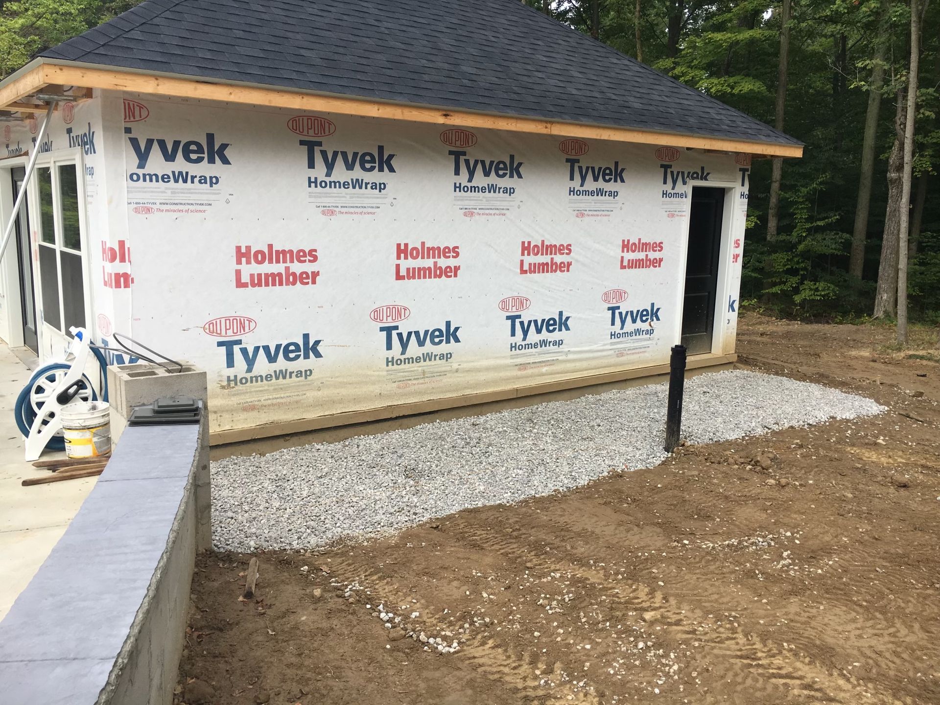 A small building under construction, wrapped in Tyvek house wrap, with a gravel path alongside it in a wooded area.