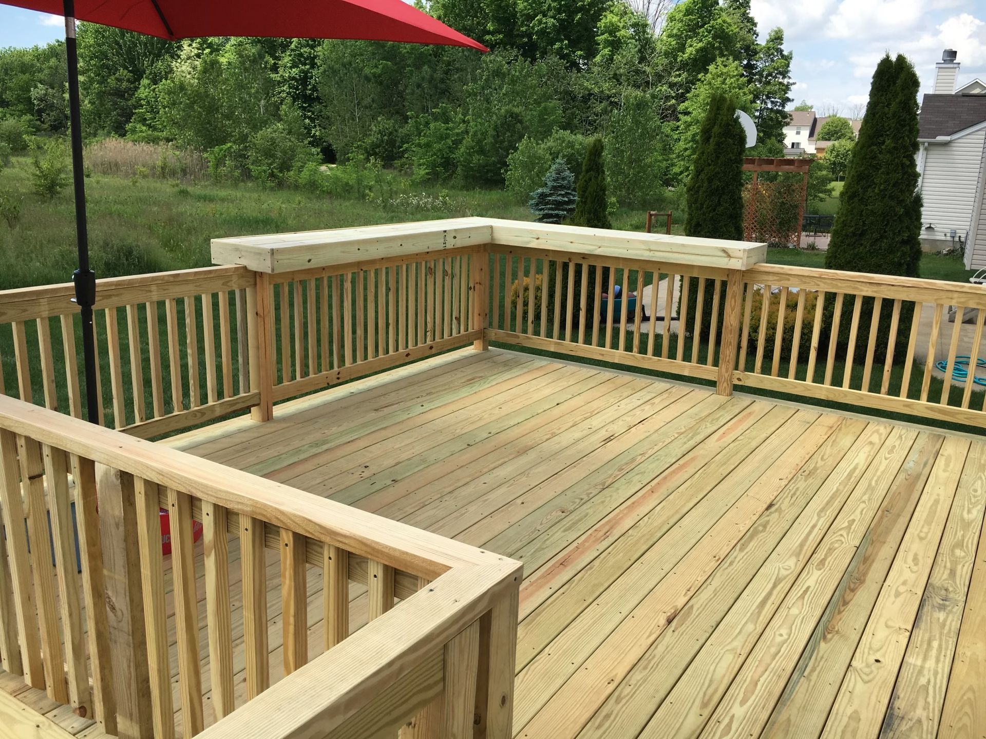 A wooden deck with railing and a bar-top edge, featuring a red umbrella, set against a green, grassy backyard.
