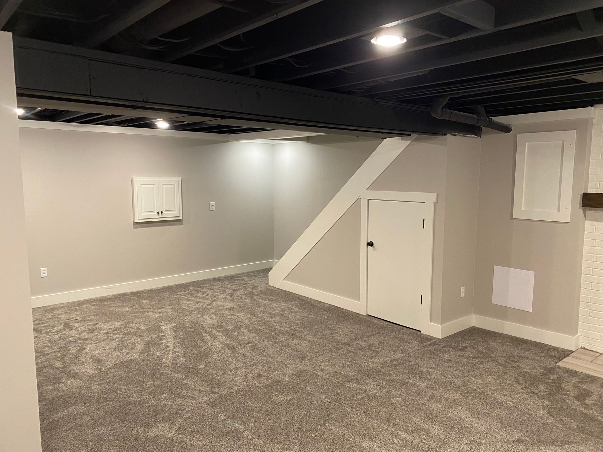 A finished basement room with gray carpet, light gray walls, a black painted exposed ceiling, and a white under-stair door.