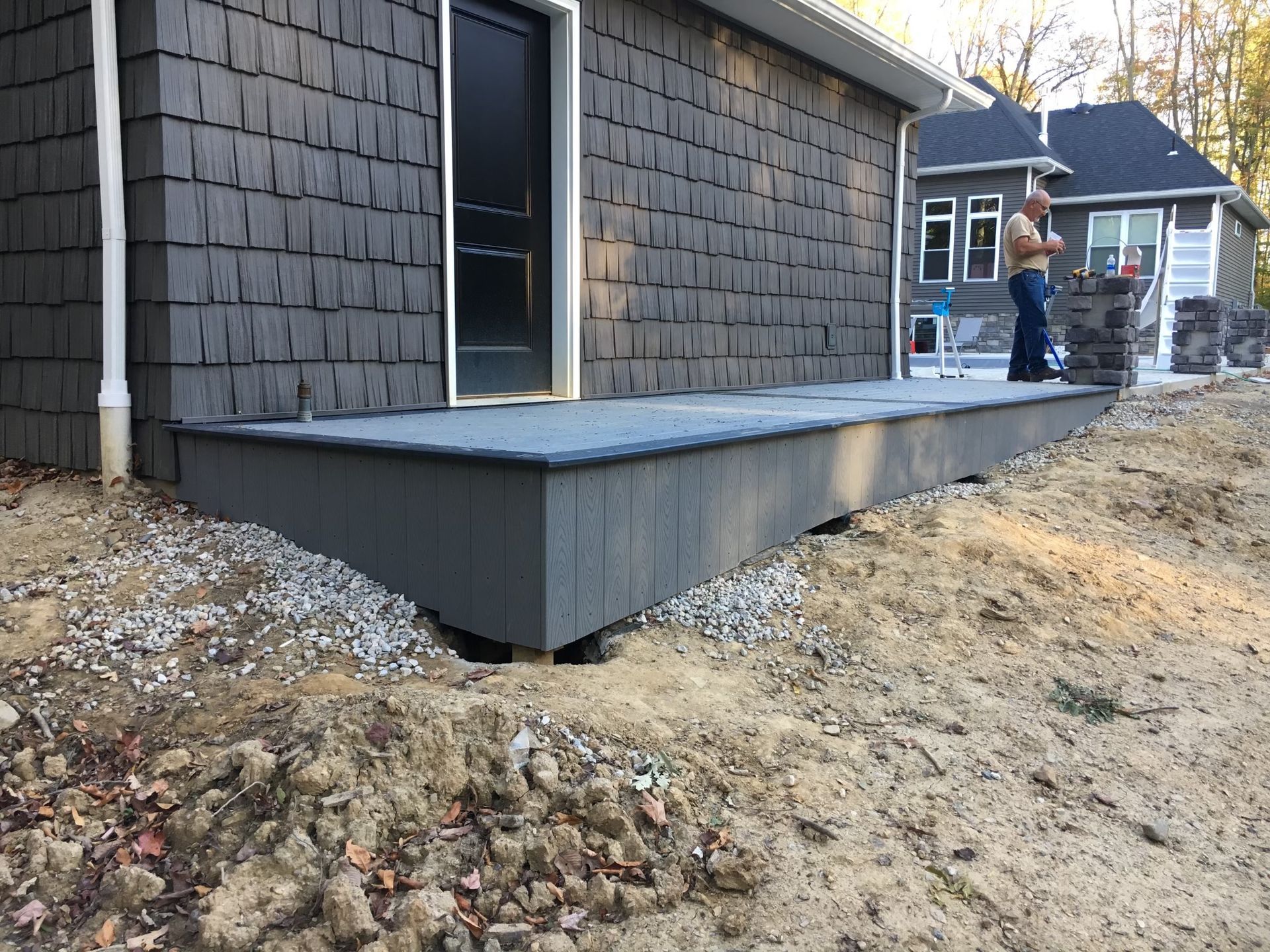 A low-profile deck with dark siding sits against a house under construction near a yard with loose dirt and gravel.