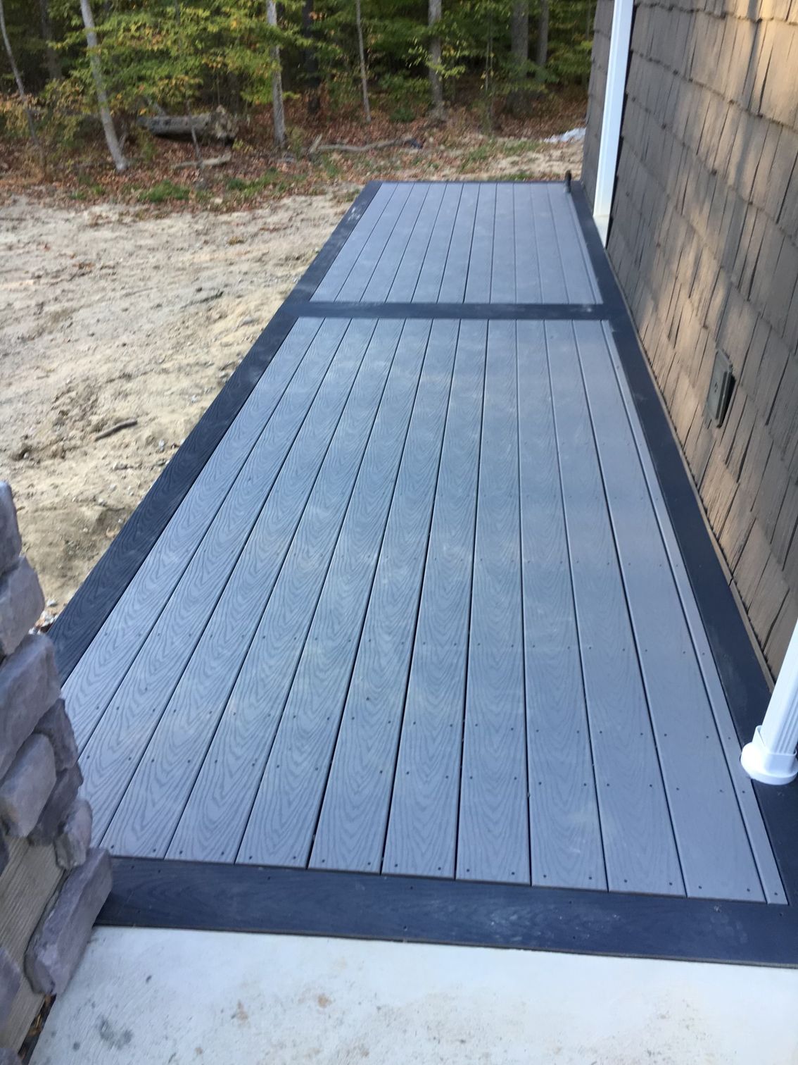 A ground-level wooden deck with gray planks and a dark-painted border next to a stone-clad house and unfinished ground.