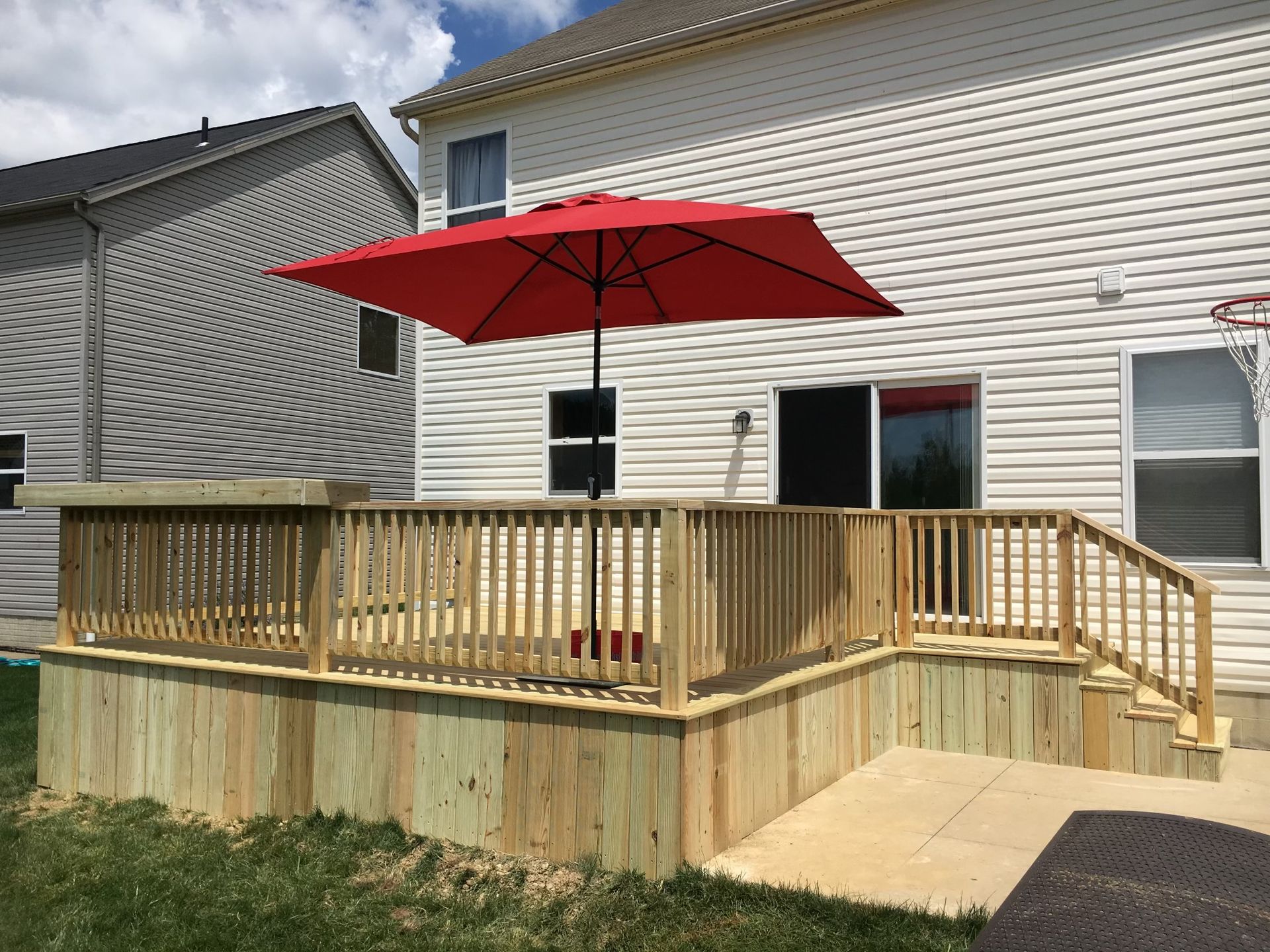 A light-colored wooden deck with a matching railing stands next to a house, featuring a large red outdoor umbrella.