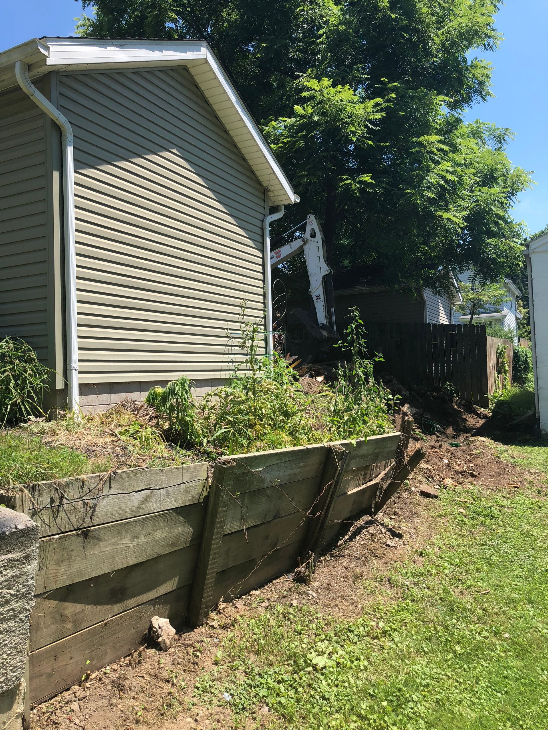 A damaged wooden retaining wall leans outward below a tan siding house with a tree and heavy machinery in the background.