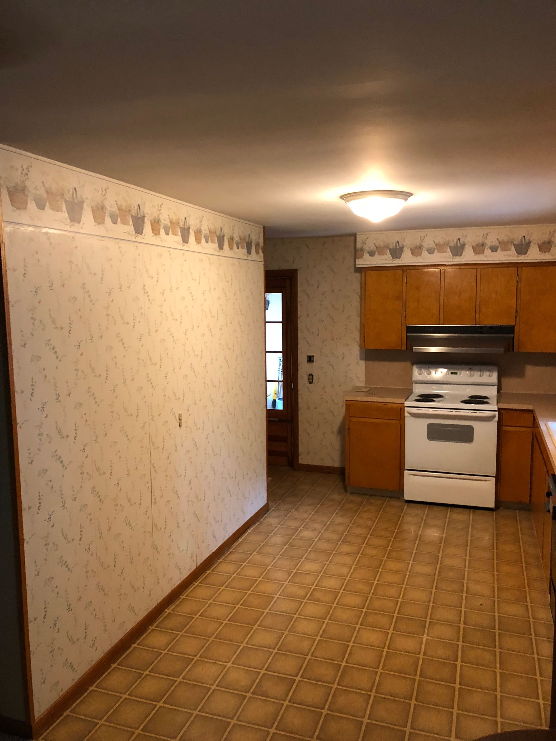 A kitchen with light-patterned wallpaper, wooden cabinets, a white stove, and brown checkered vinyl flooring.