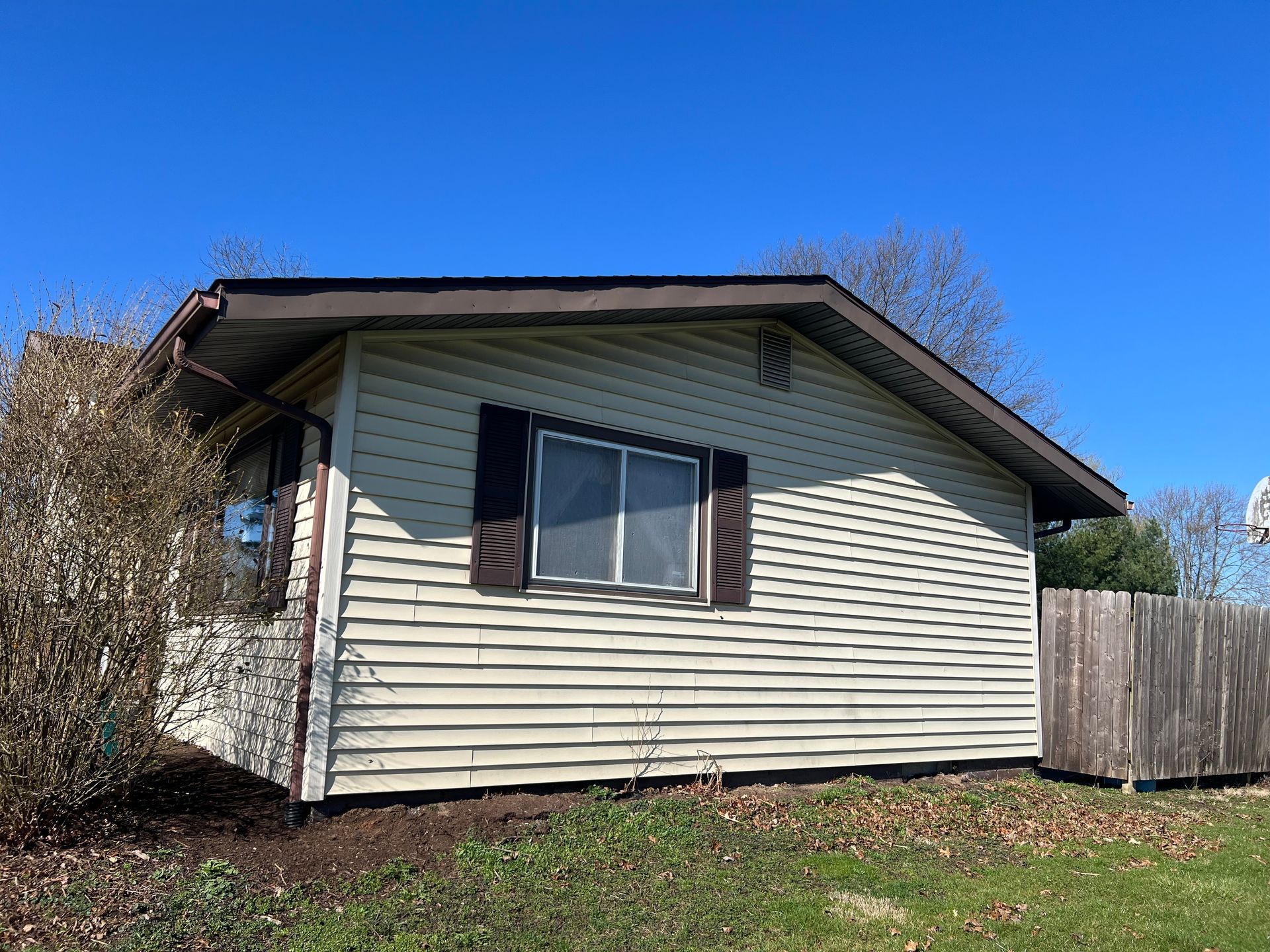 Single-story house with cream-colored horizontal siding, a dark roof, and a single window with dark shutters.