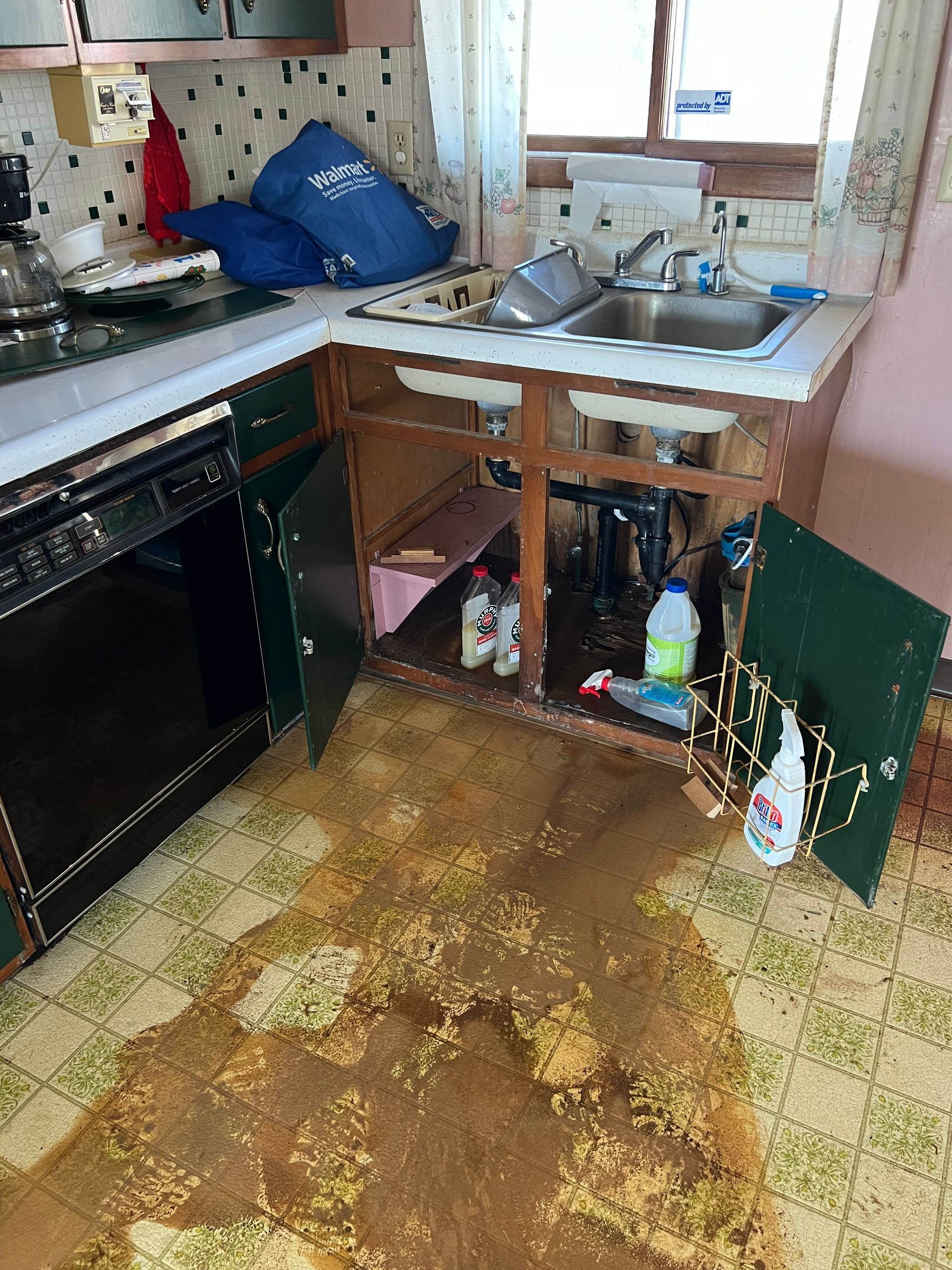 A kitchen sink with open cabinets leaking a large, dark-colored puddle onto the linoleum floor.
