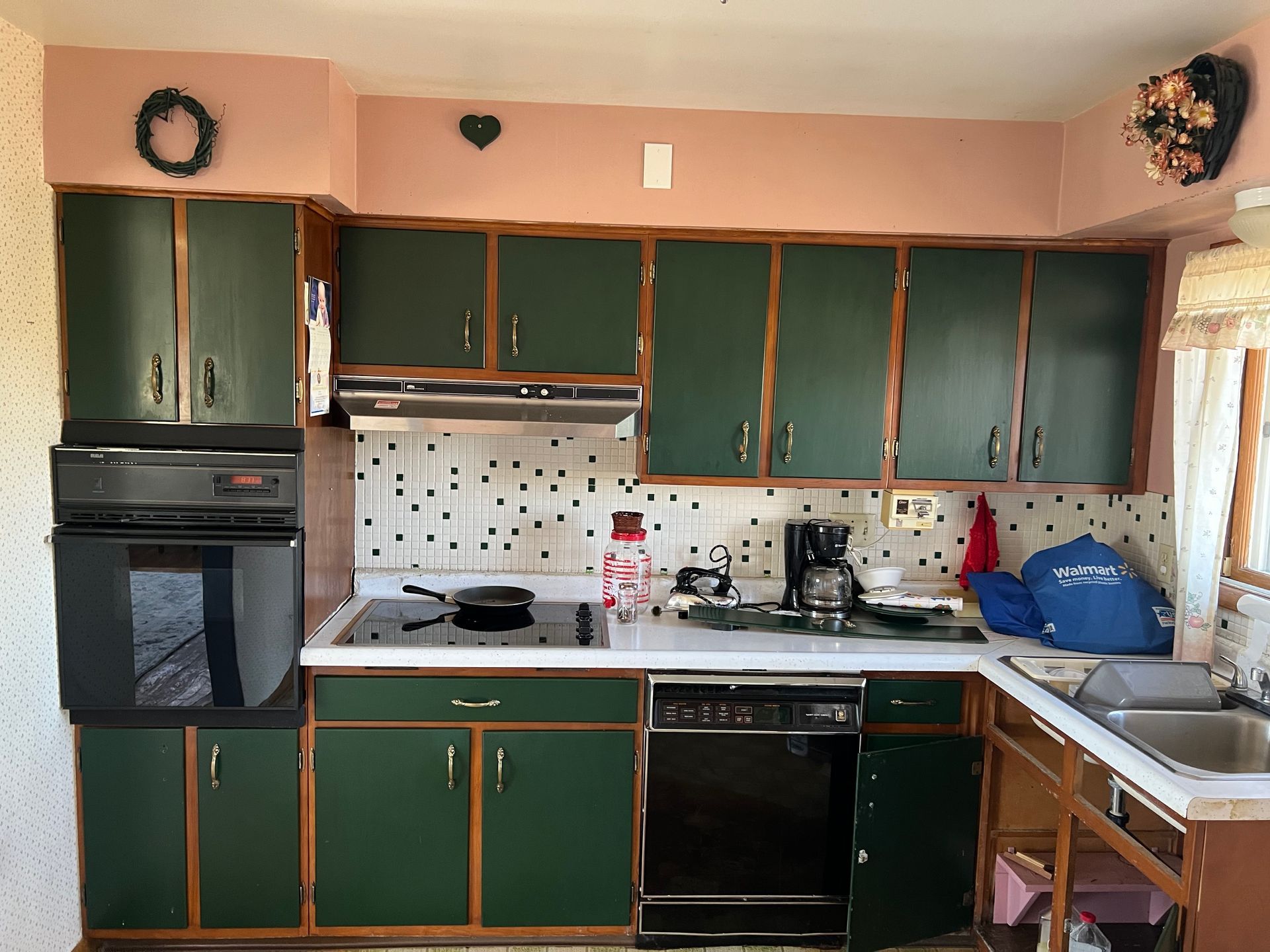 A kitchen with dark green cabinets, a wall-mounted oven, a glass stovetop, a white tiled backsplash, and a sink.