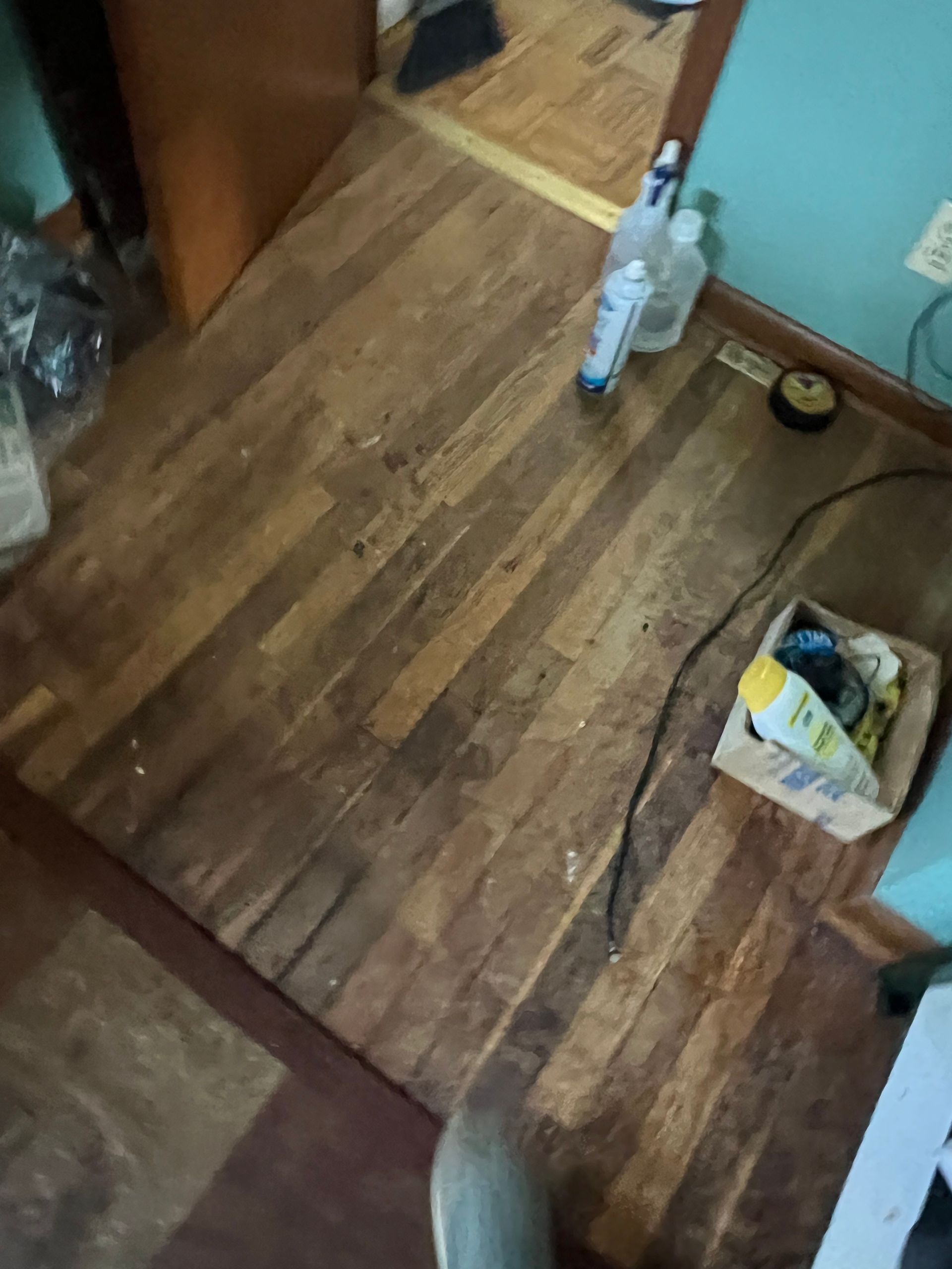 A slightly cluttered room with wood-patterned flooring, a doorway, cleaning supplies, and a box of items on the floor.