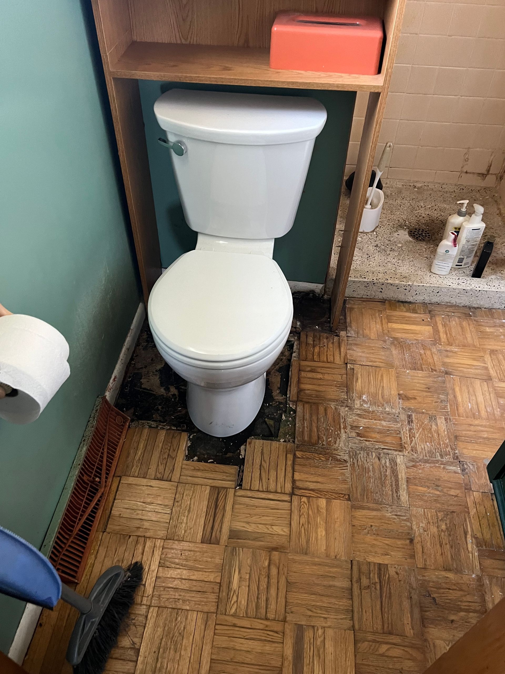 A white toilet in a bathroom with parquet wood flooring and a wooden over-the-toilet storage unit holding a tissue box.