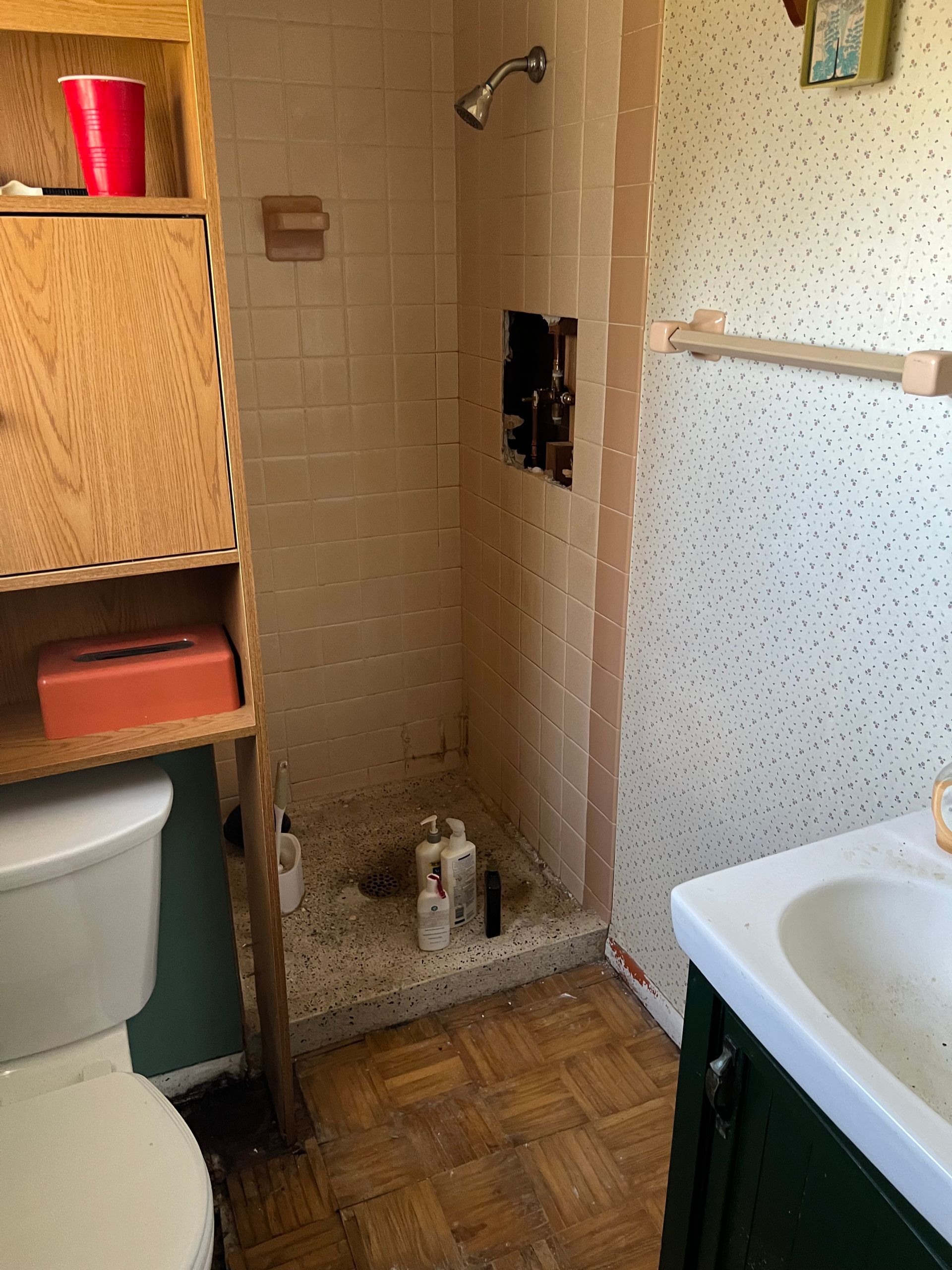 A small bathroom with a tile shower stall missing its handle, a toilet, a wooden shelf, and a sink.