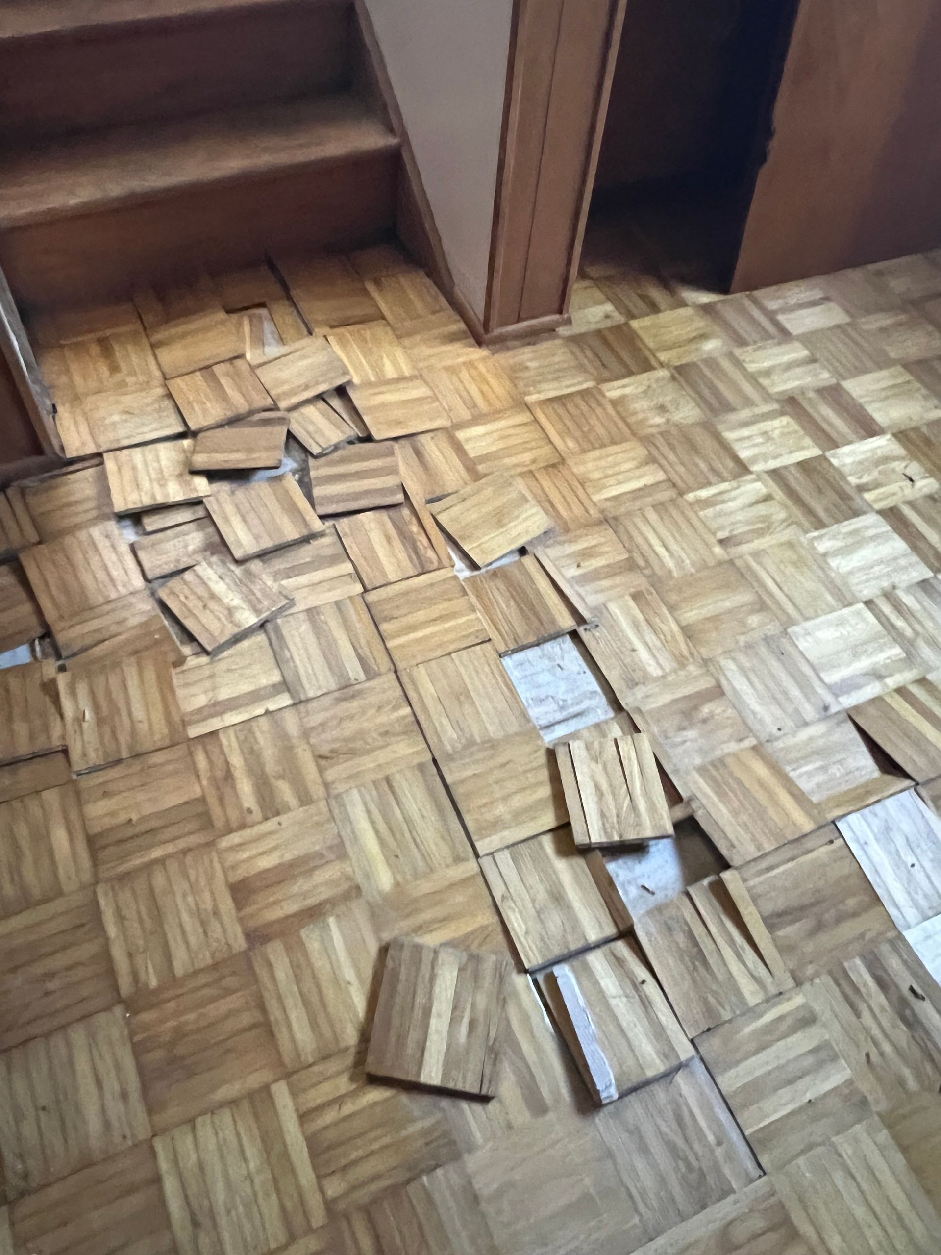 Damaged parquet wood flooring with several loose tiles scattered on the floor near a staircase.