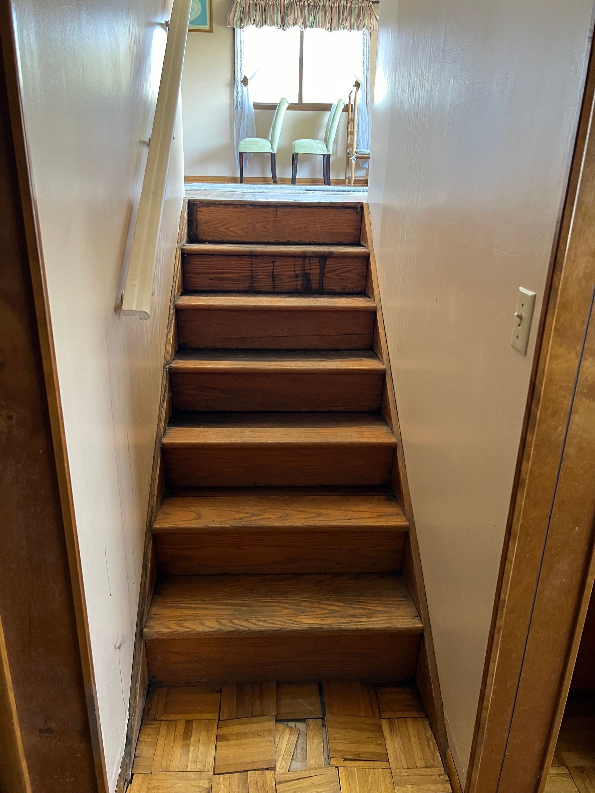 Wooden staircase between two white walls, leading to a sunlit room with dining chairs visible at the top.