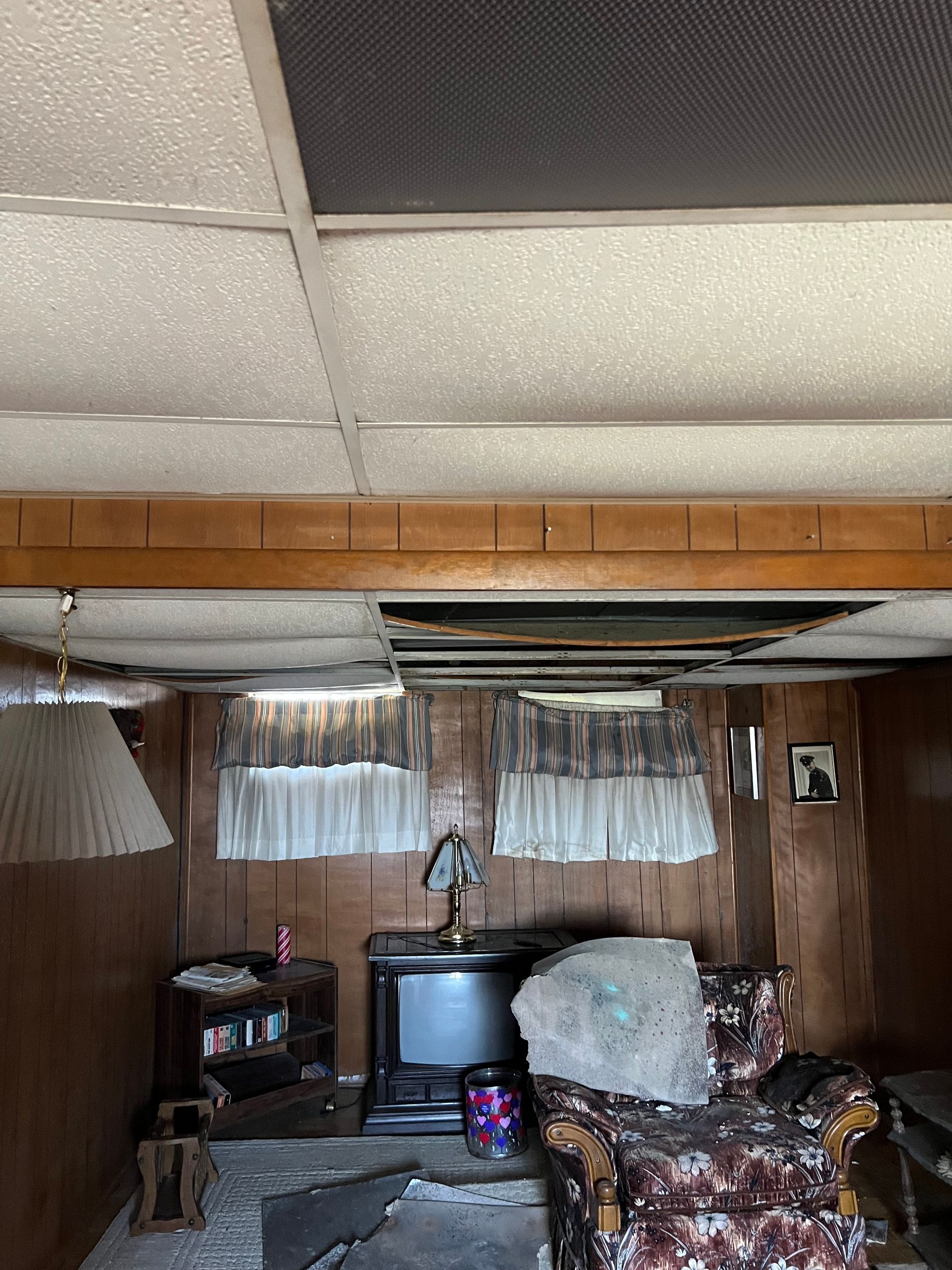 A dimly lit room with wood-paneled walls, a drop ceiling with missing tiles, a small TV, and a floral armchair.