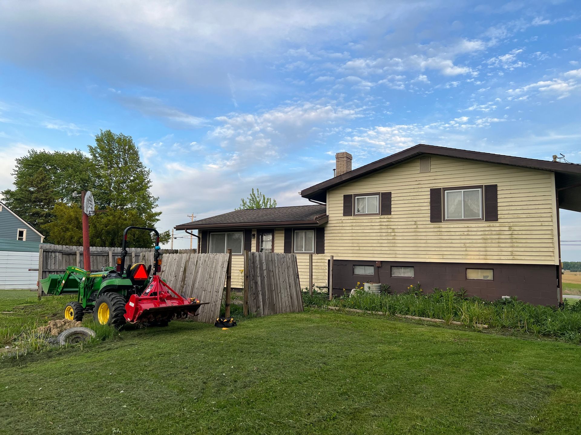 A green John Deere tractor with a red attachment parked in the yard of a yellow house with a dark brown foundation.