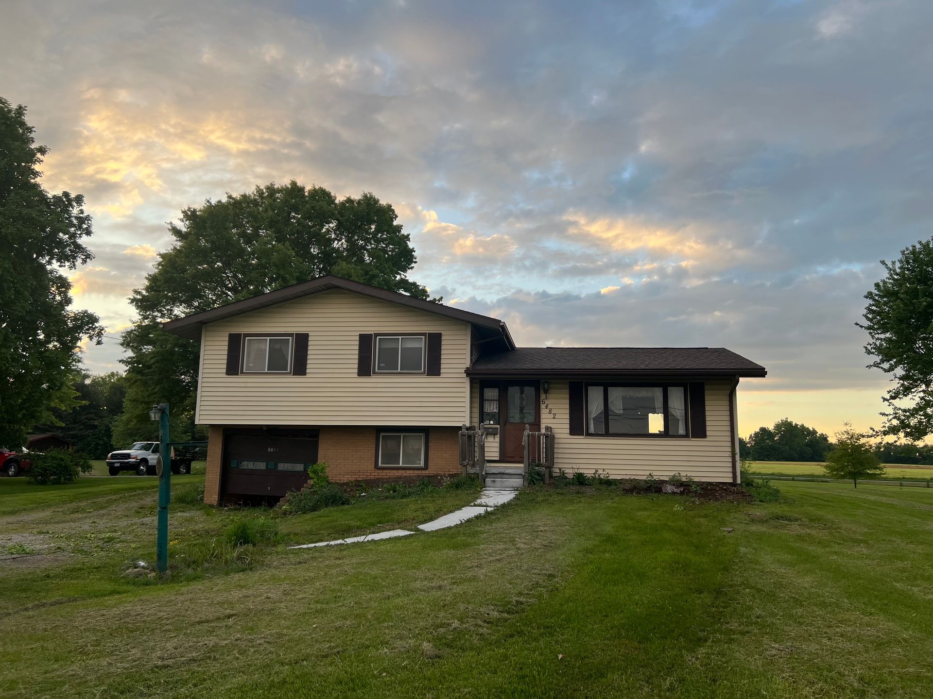 A two-story tan suburban house with a brown roof sits on a grassy lot under a sunset sky.