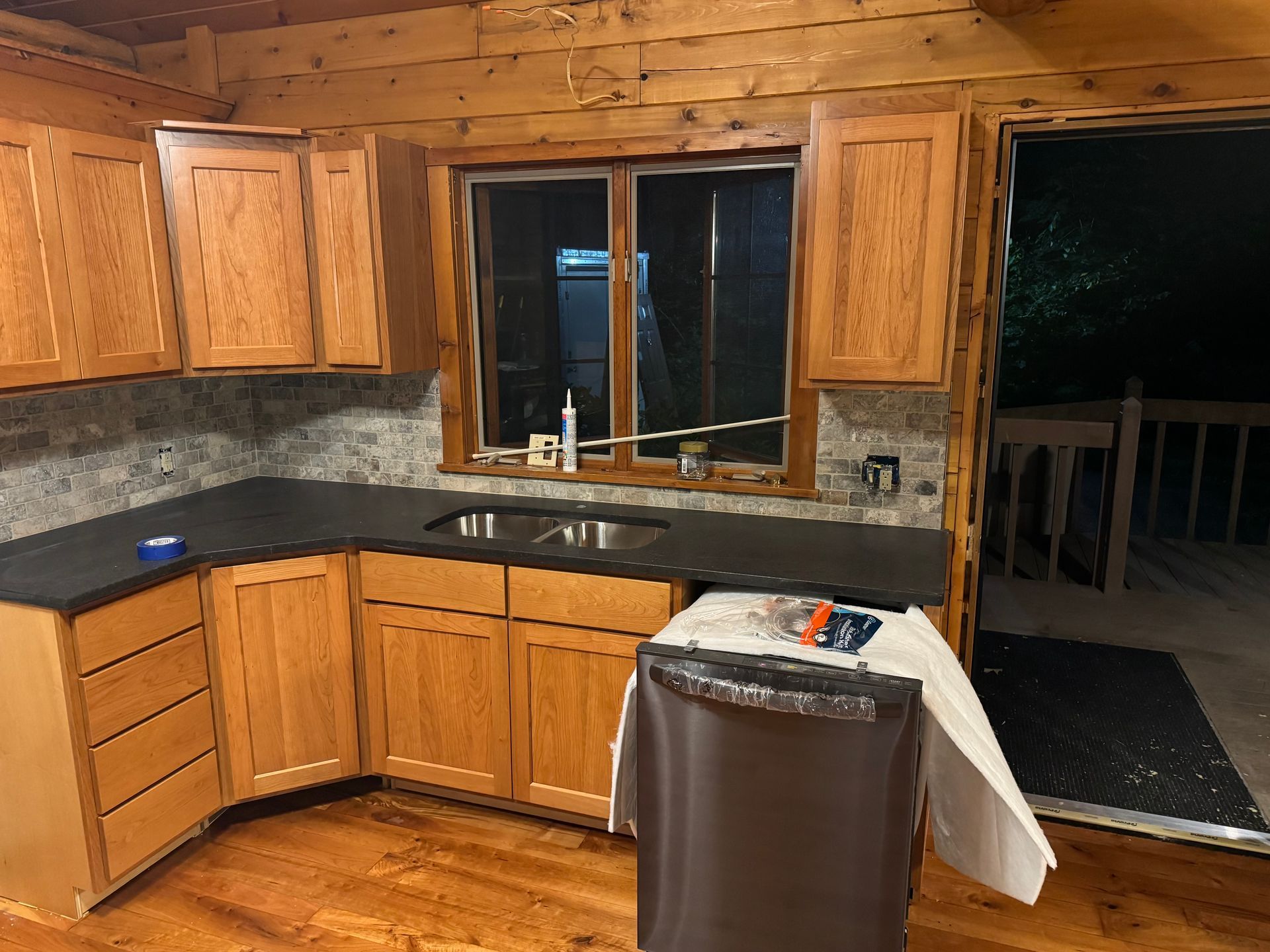 A rustic kitchen with wood-paneled walls, oak cabinets, dark countertops, a sink, and a dishwasher under renovation.