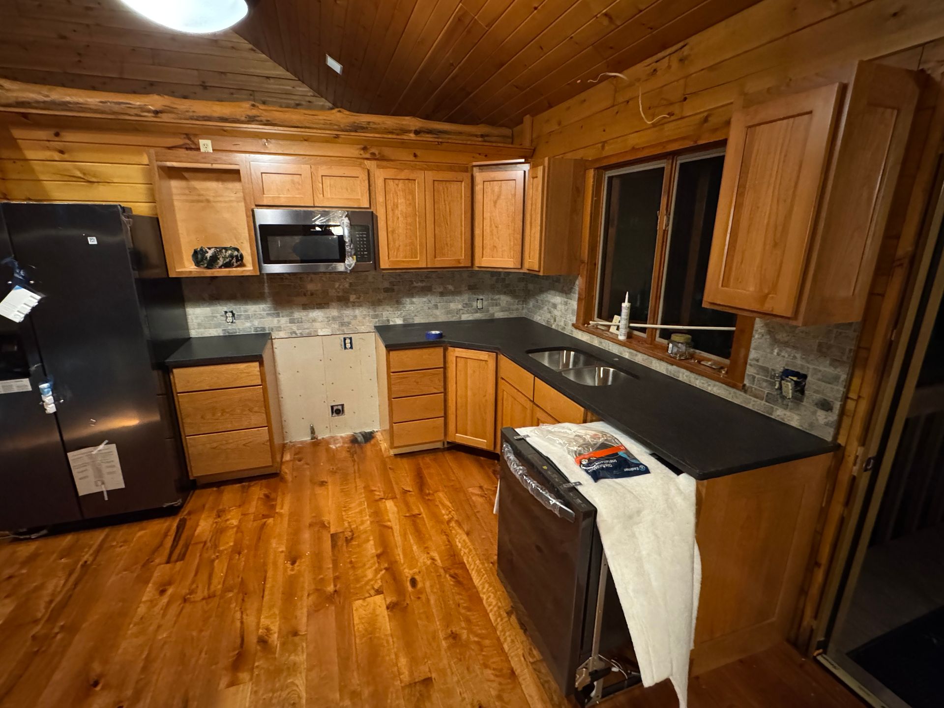 A rustic kitchen with wood walls, cabinets, dark countertops, and stainless steel appliances in a log home.