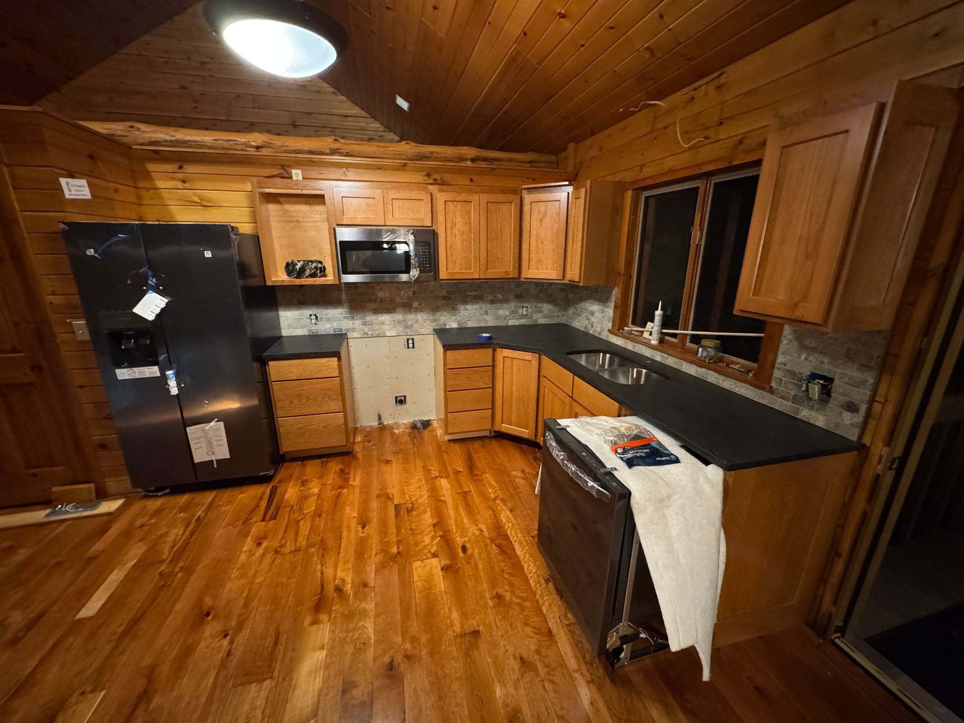 A kitchen with wooden walls, cabinets, and flooring, black countertops, a dark refrigerator, and a dishwasher with a towel.