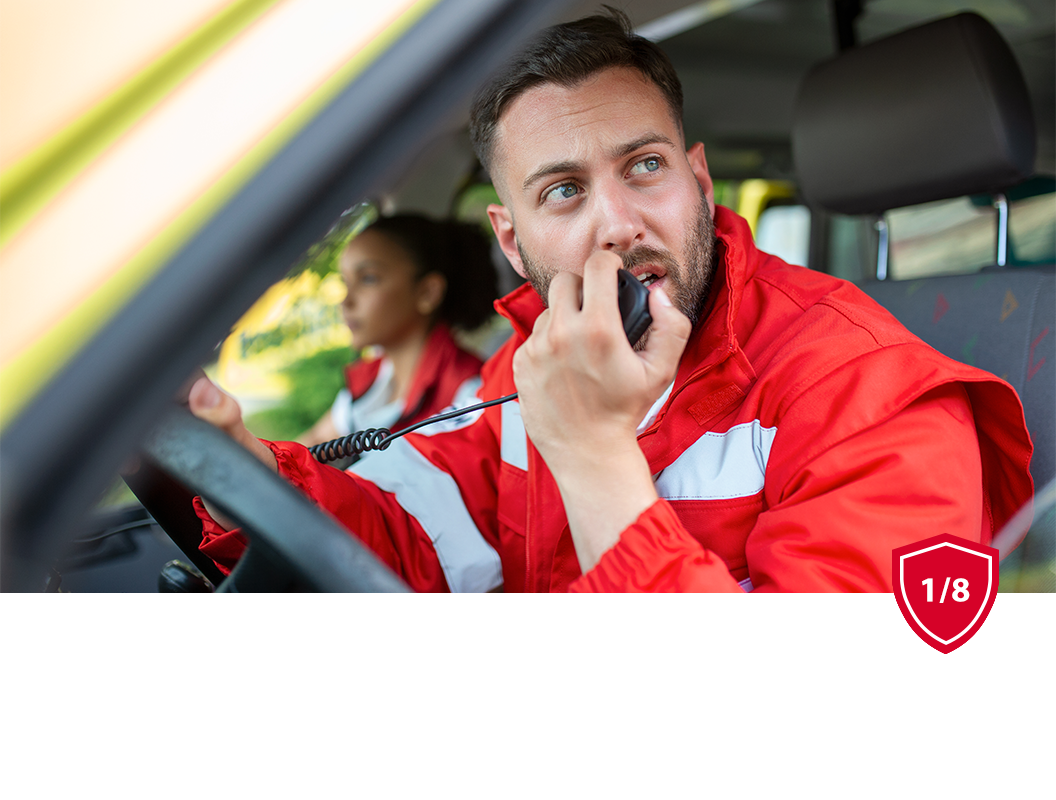 Two emergency medical responders in red uniform jackets sit in the front of a vehicle, with the driver using a radio mic.