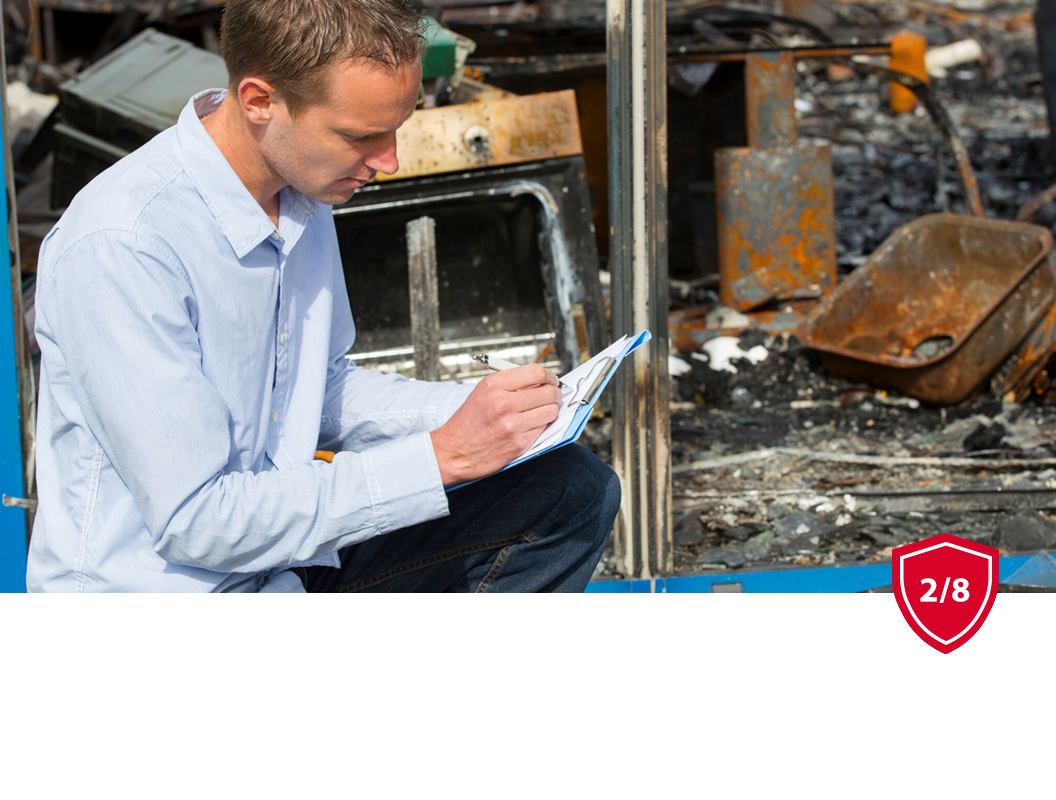 A person in a light blue shirt kneels to write on a clipboard in front of debris from a fire-damaged building.