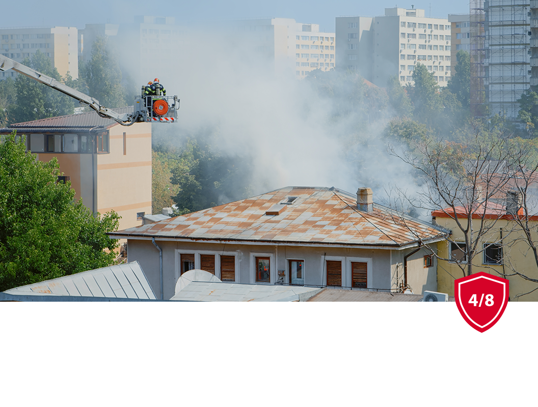 A firefighter in an aerial ladder bucket sprays water onto a smoke-filled building roof near residential apartment blocks.