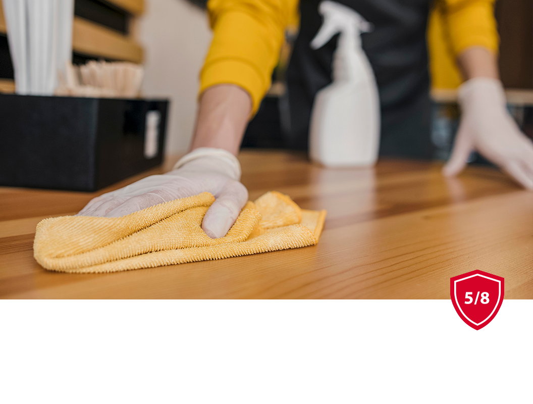 A person wearing gloves wipes a wooden counter with a yellow cloth, with a spray bottle in the background.