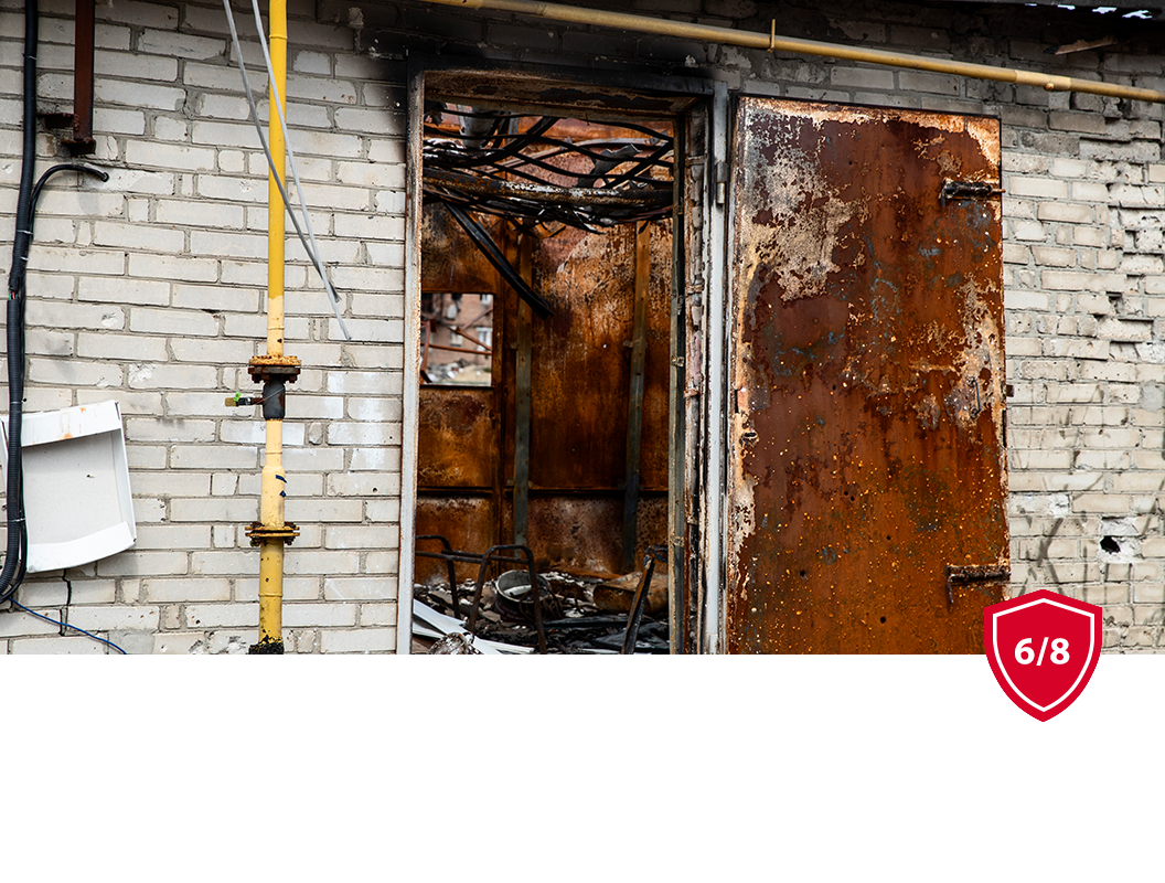 A view of a damaged, burnt building interior through an open metal door set into a weathered white brick wall.