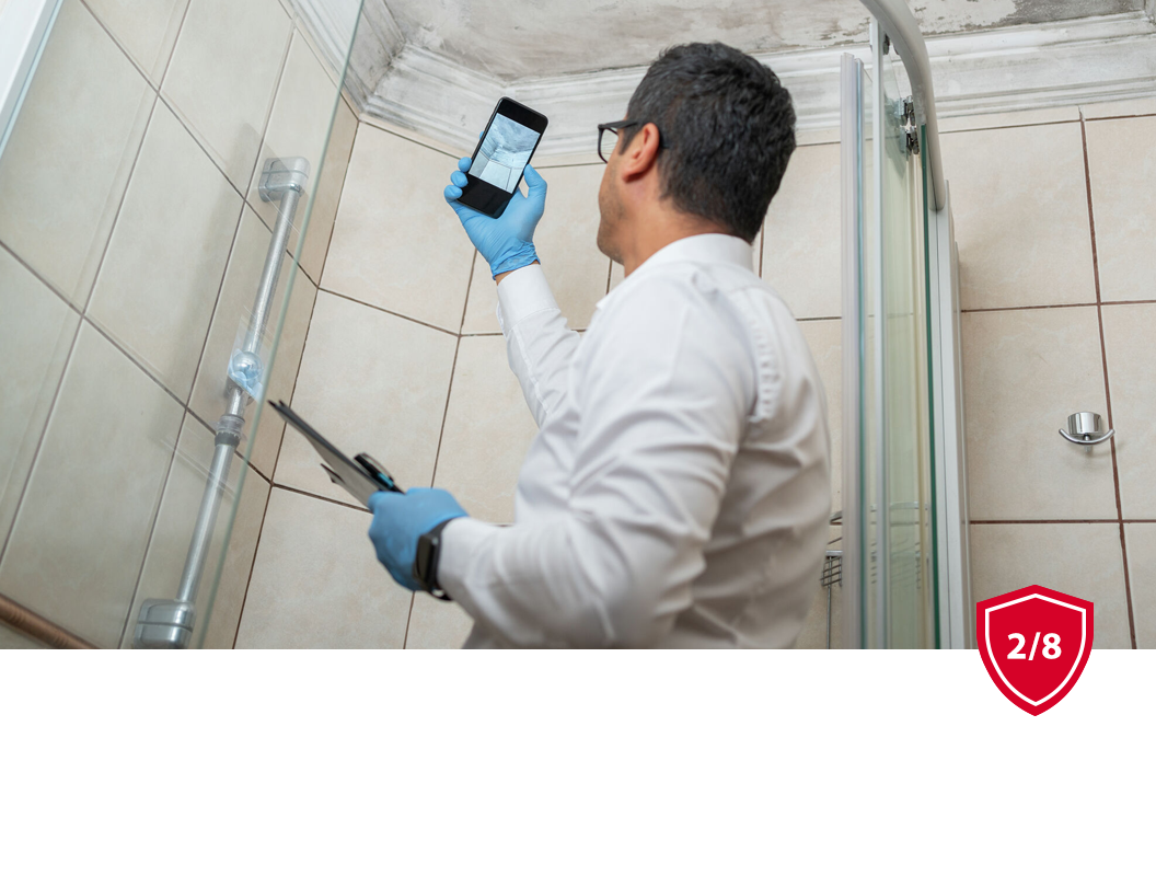 A person in a white shirt and blue gloves holds a phone up to photograph a tiled shower area while holding a clipboard.