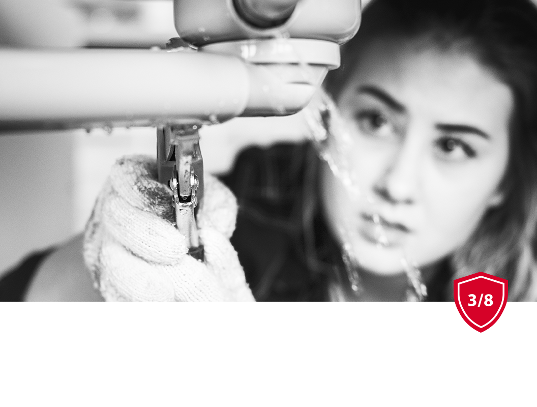 A focused person wears gloves while using pliers to tighten or adjust plumbing pipes under a sink with water spraying out.