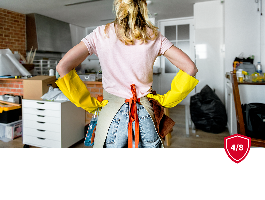 A person wearing yellow rubber cleaning gloves and an apron stands with hands on their hips in a cluttered room.