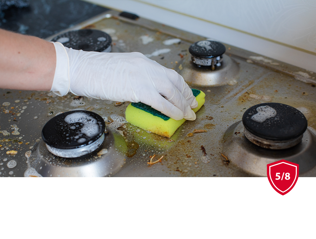 A person wearing a white glove scrubs a greasy kitchen stovetop with a yellow and green sponge.