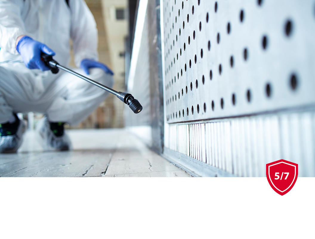 A person in a white hazmat suit and blue gloves uses a spray wand to sanitize a metal wall with perforated holes.