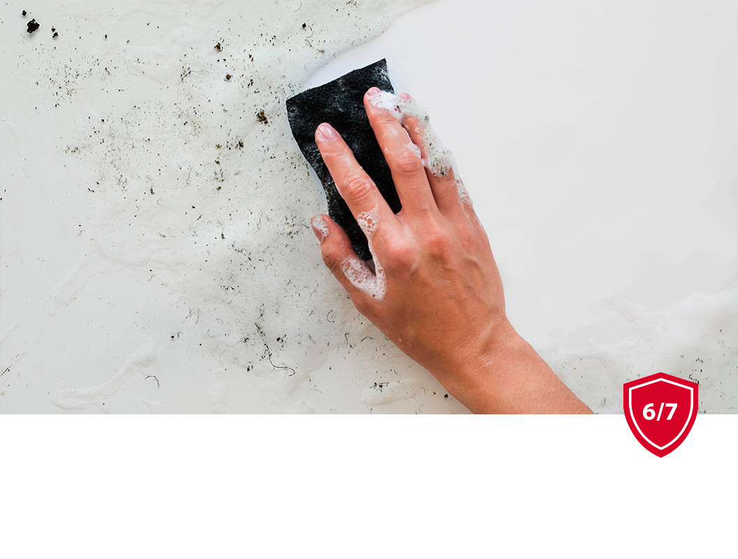 A hand uses a dark cleaning sponge to scrub dark grime off a white surface.